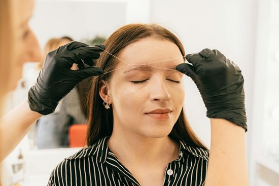 A woman having her eyebrows measured with thread by someone wearing black gloves in a beauty salon.