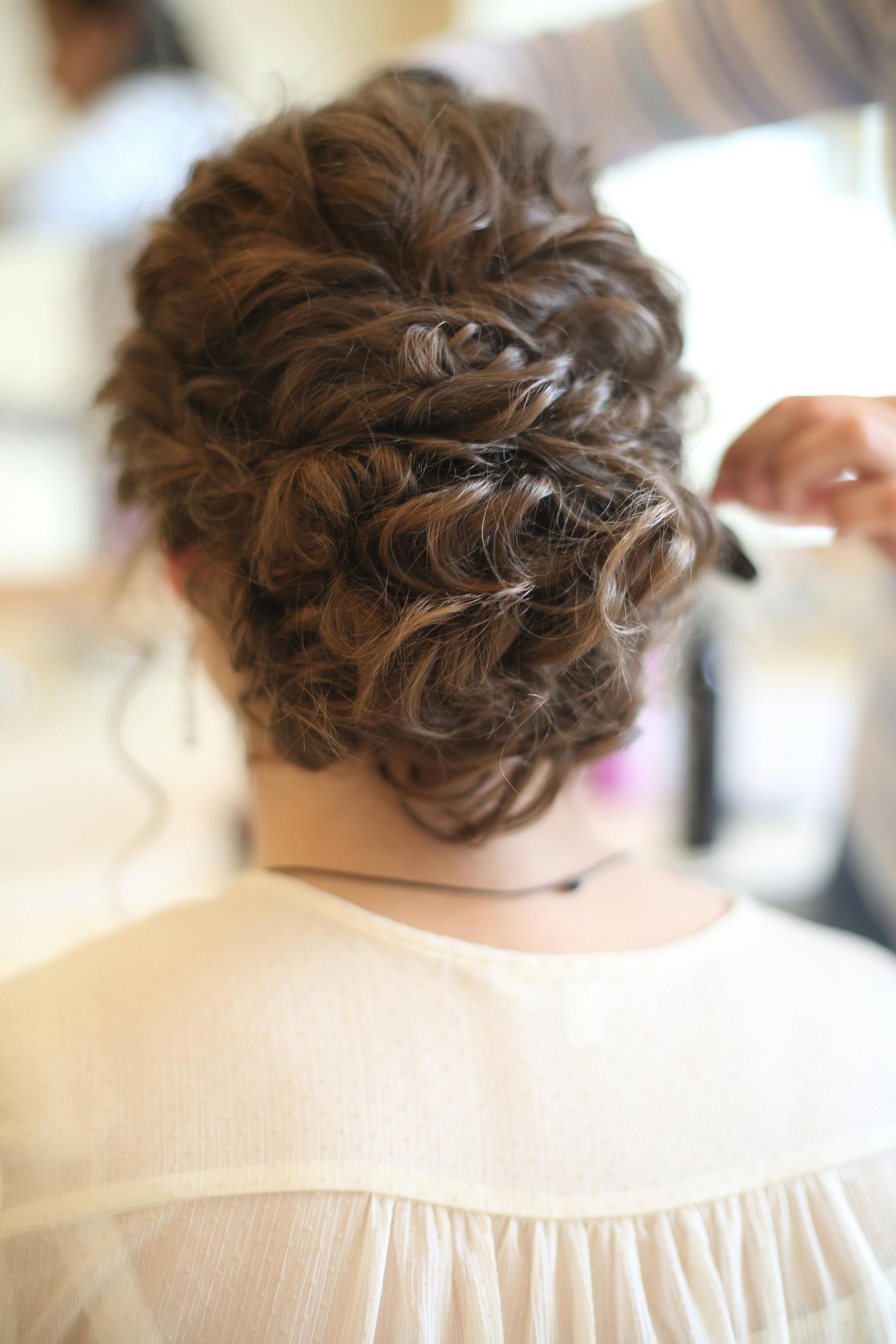 Woman with curly brown hair styled in an updo, being styled by a person in a light-colored top.