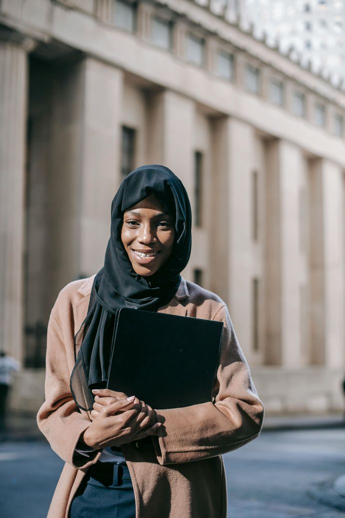 A woman wearing a hijab is holding a laptop in front of a building.