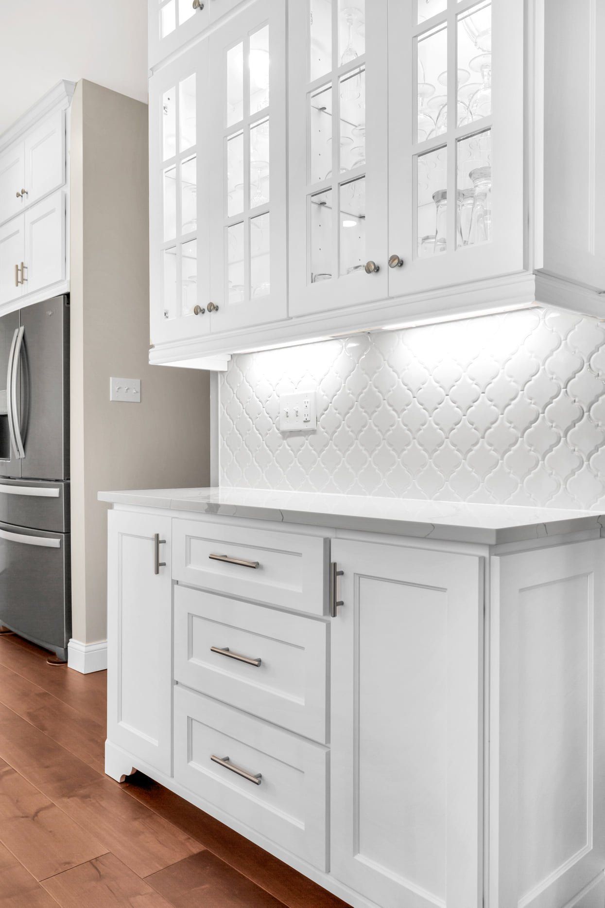 White kitchen cabinets with under-cabinet lighting, gray tile backsplash, and wood floor