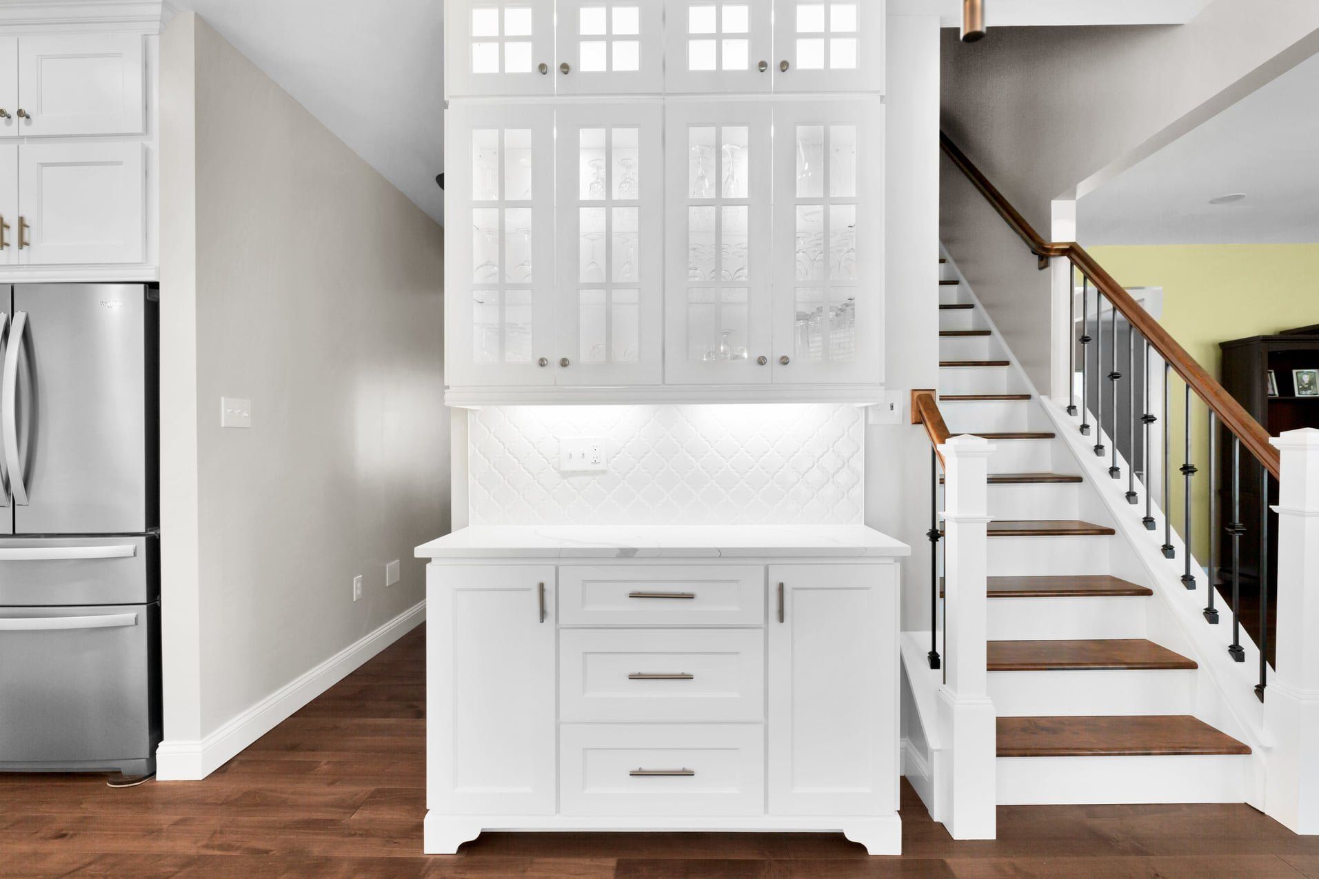 Bright white kitchen nook with built-in cabinets, stainless fridge, wood floors, and staircase nearby