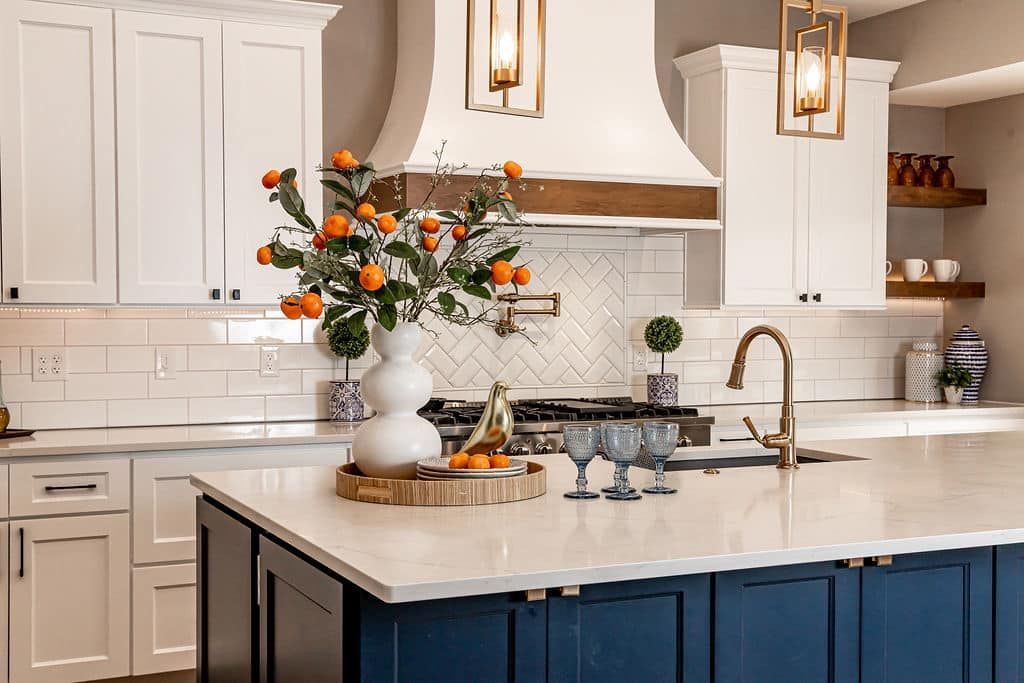 Bright white kitchen with navy island, marble counters, brass lights, and orange flowers in a vase