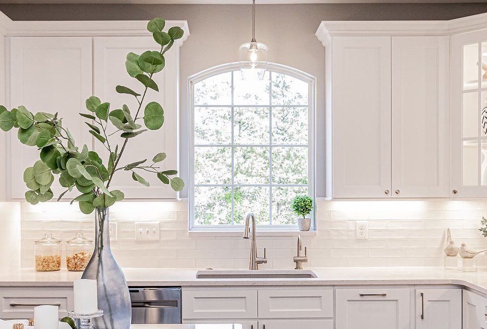 Bright white kitchen with a tall leafy branch in a vase by a window over the sink.