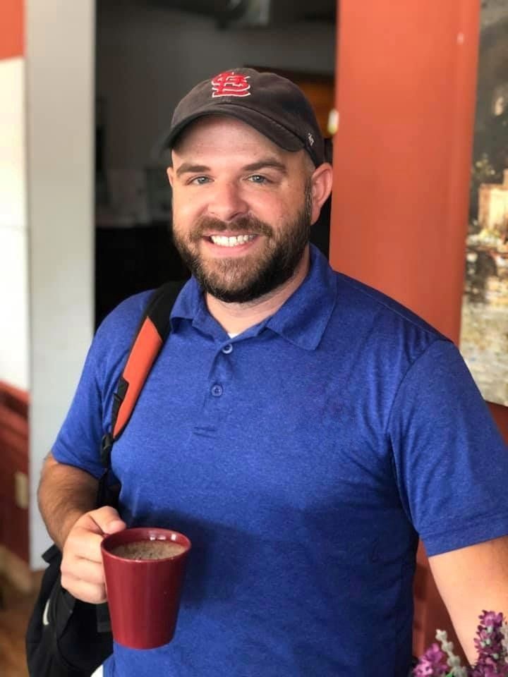 Smiling man in a blue shirt and black cap holding a red mug indoors