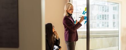 Een vrouw schrijft op een whiteboard terwijl een andere vrouw toekijkt, als illustratie van het ontwikkelen van analytische vaardigheden.