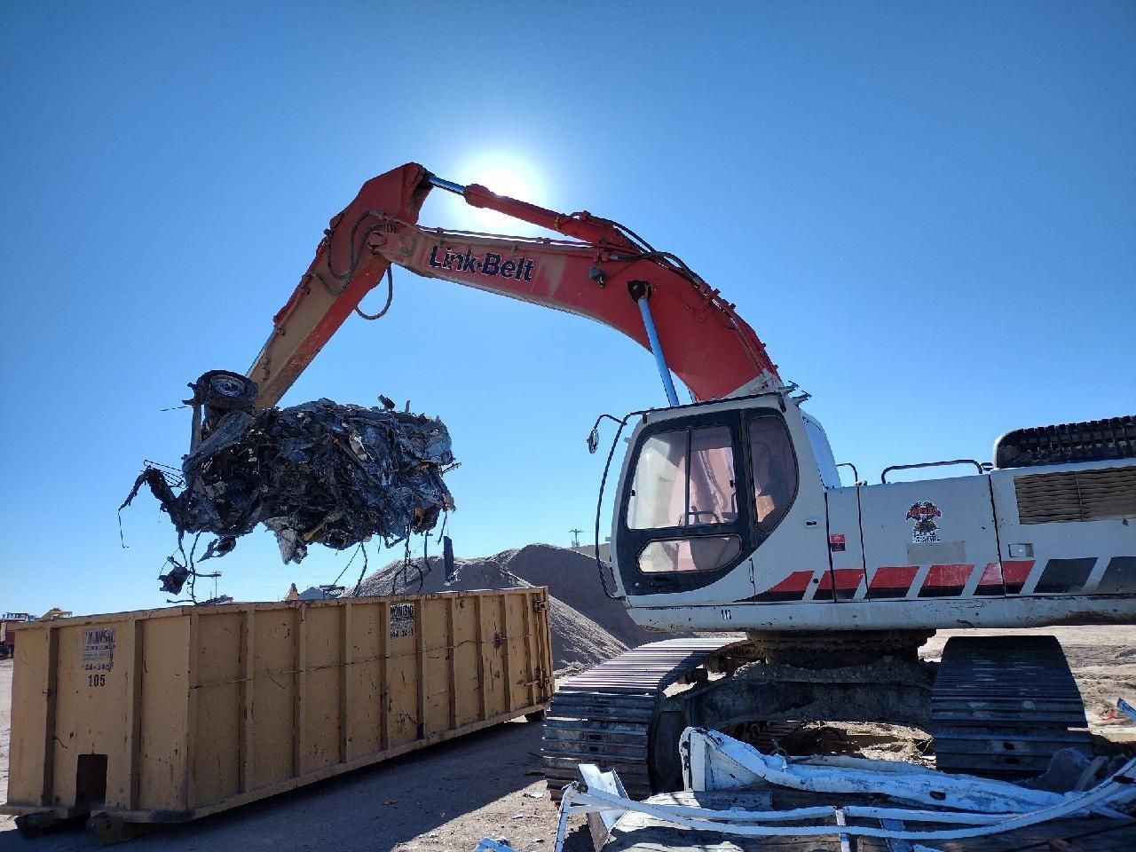 A large excavator is loading a dumpster with scrap metal