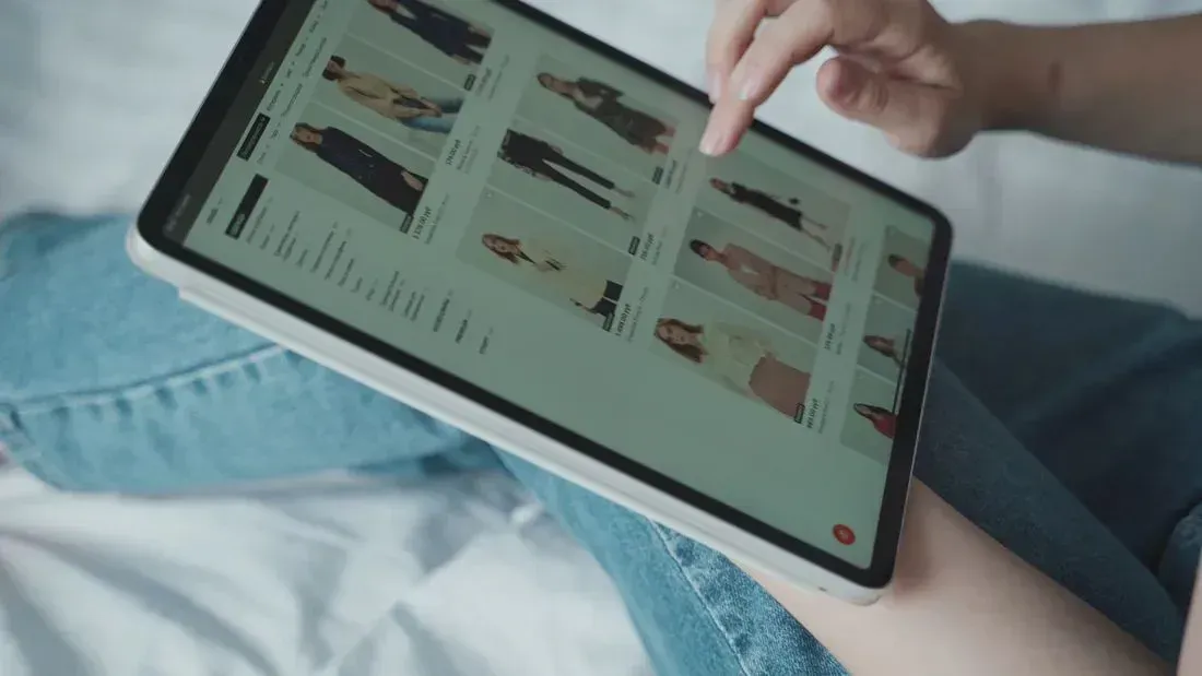 Person using a tablet on a bed, browsing a shopping page with denim jeans visible.