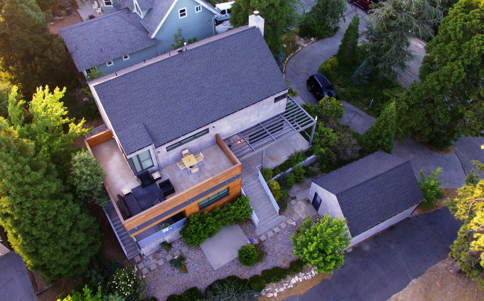 Drone Aerial view of a suburban house with a gray roof, wooden deck, driveway, and surrounding trees and shrubs.