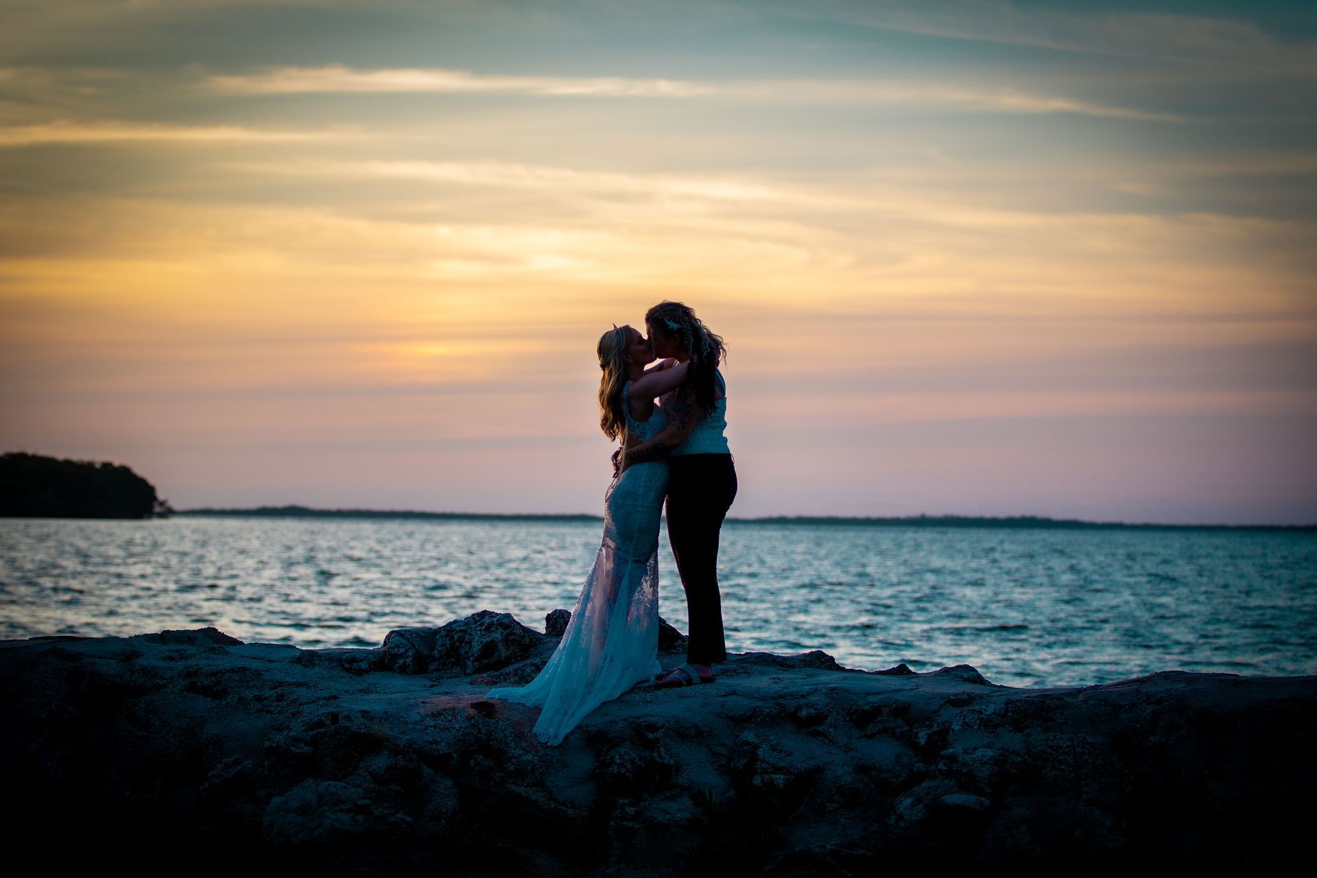 Drone Couple embracing on a rocky shore at sunset, with ocean and pastel sky behind them
