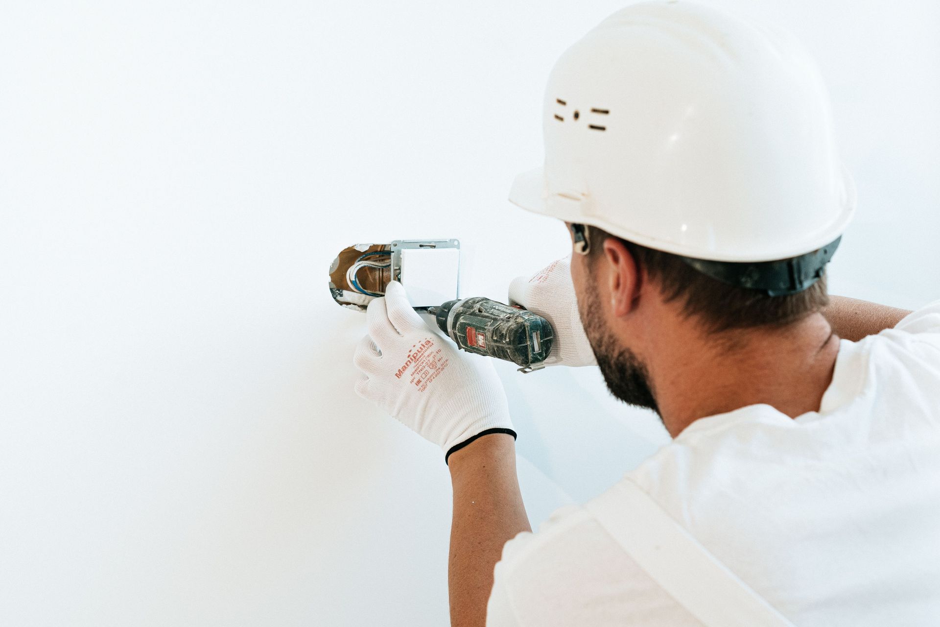 Worker installing a wall fixture with a drill, wearing a white hard hat and gloves