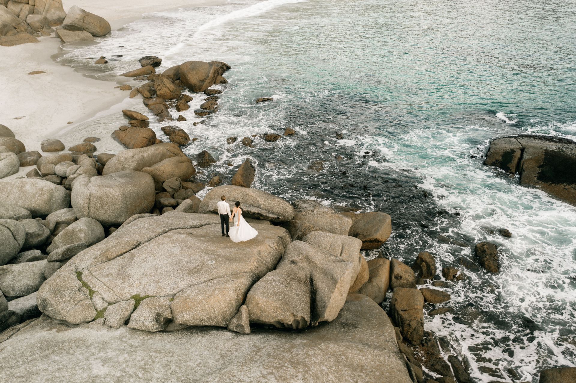 Drone Person in white sitting on large rocks by a foamy shoreline with clear water.
