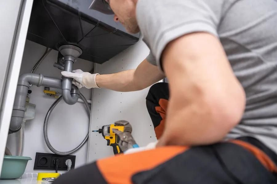 Plumber working under a sink, using a drill on pipes, wearing gloves and orange pants.