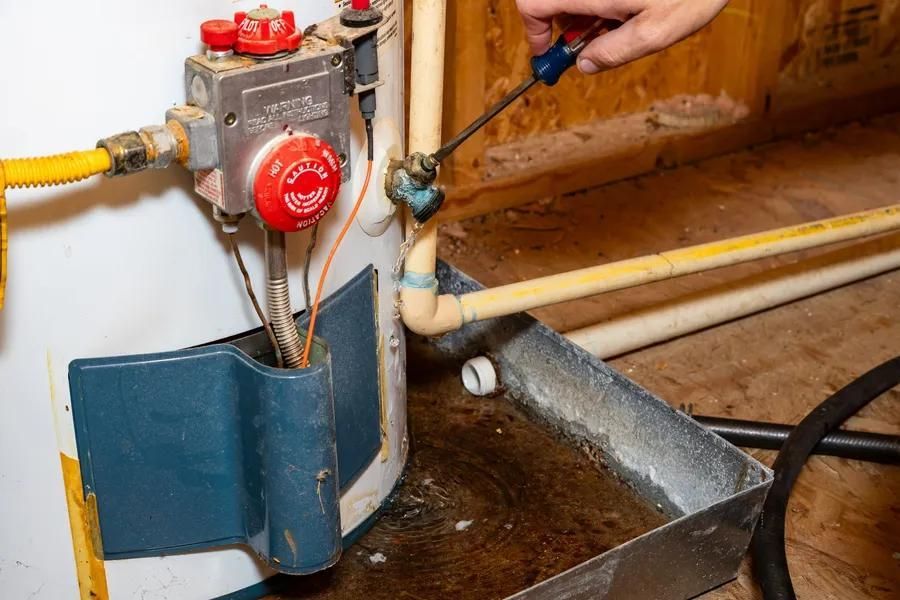 Person using a screwdriver on a leaky water heater pipe, water in a tray below.