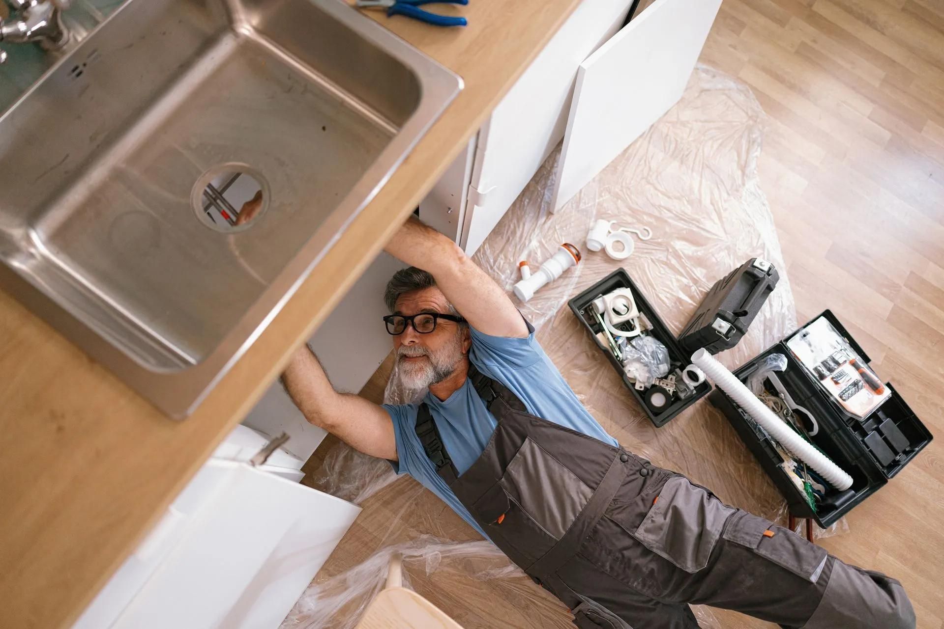 Plumber works under a kitchen sink, surrounded by tools and parts, on a light wood floor.