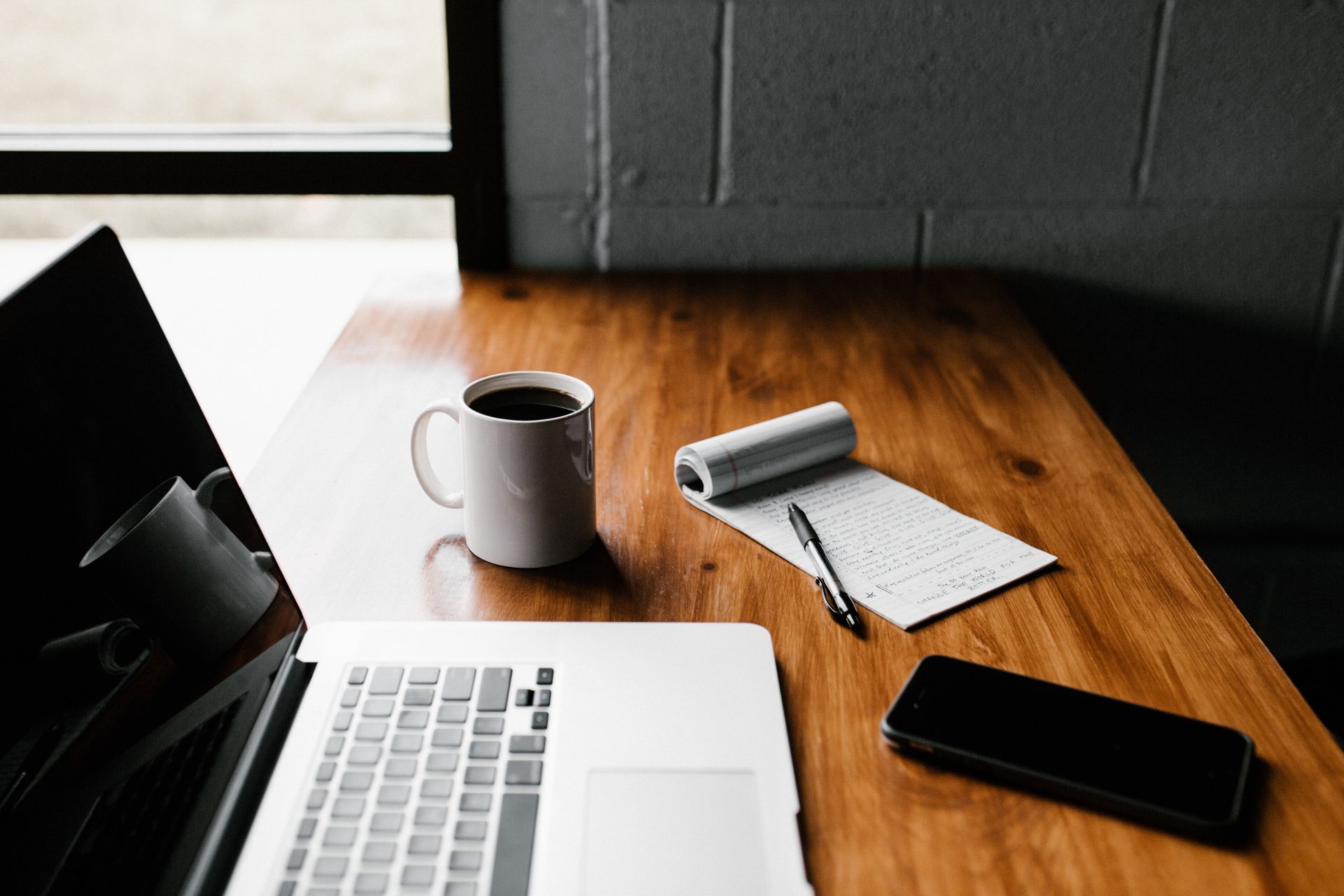 Laptop, coffee mug, notebook, and smartphone on a wooden desk by a window.