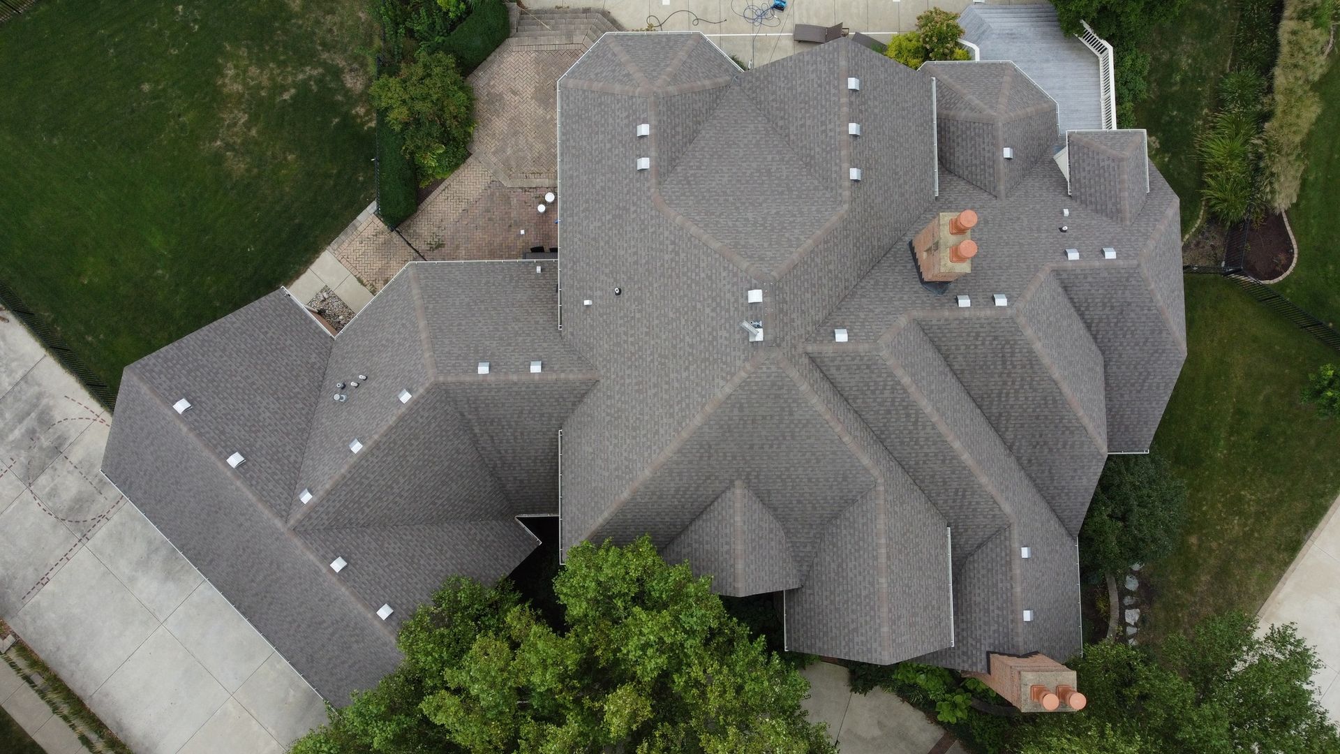 An aerial view of a large, shingled residential roof with multiple peaks, chimneys, and surrounding trees and driveways.