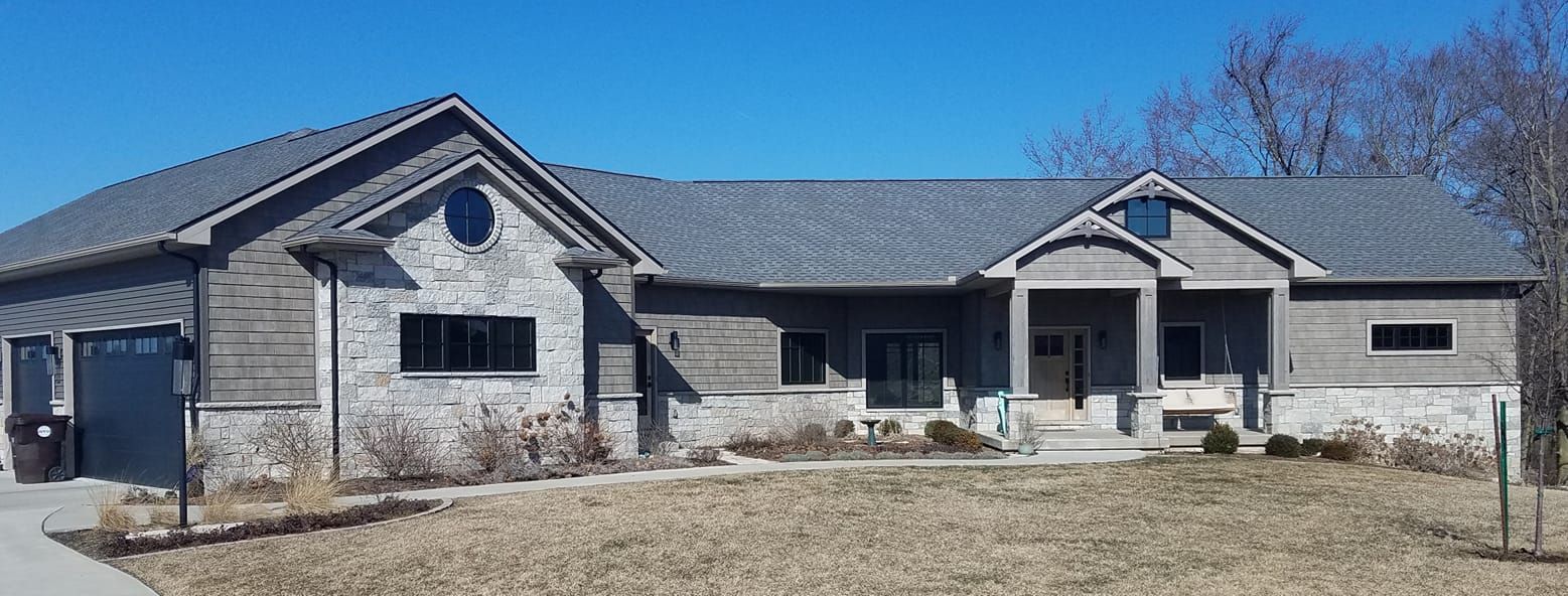 A single-story, grey-sided home with stone accents, a dark shingled roof, a front porch, and a driveway, under a blue sky.