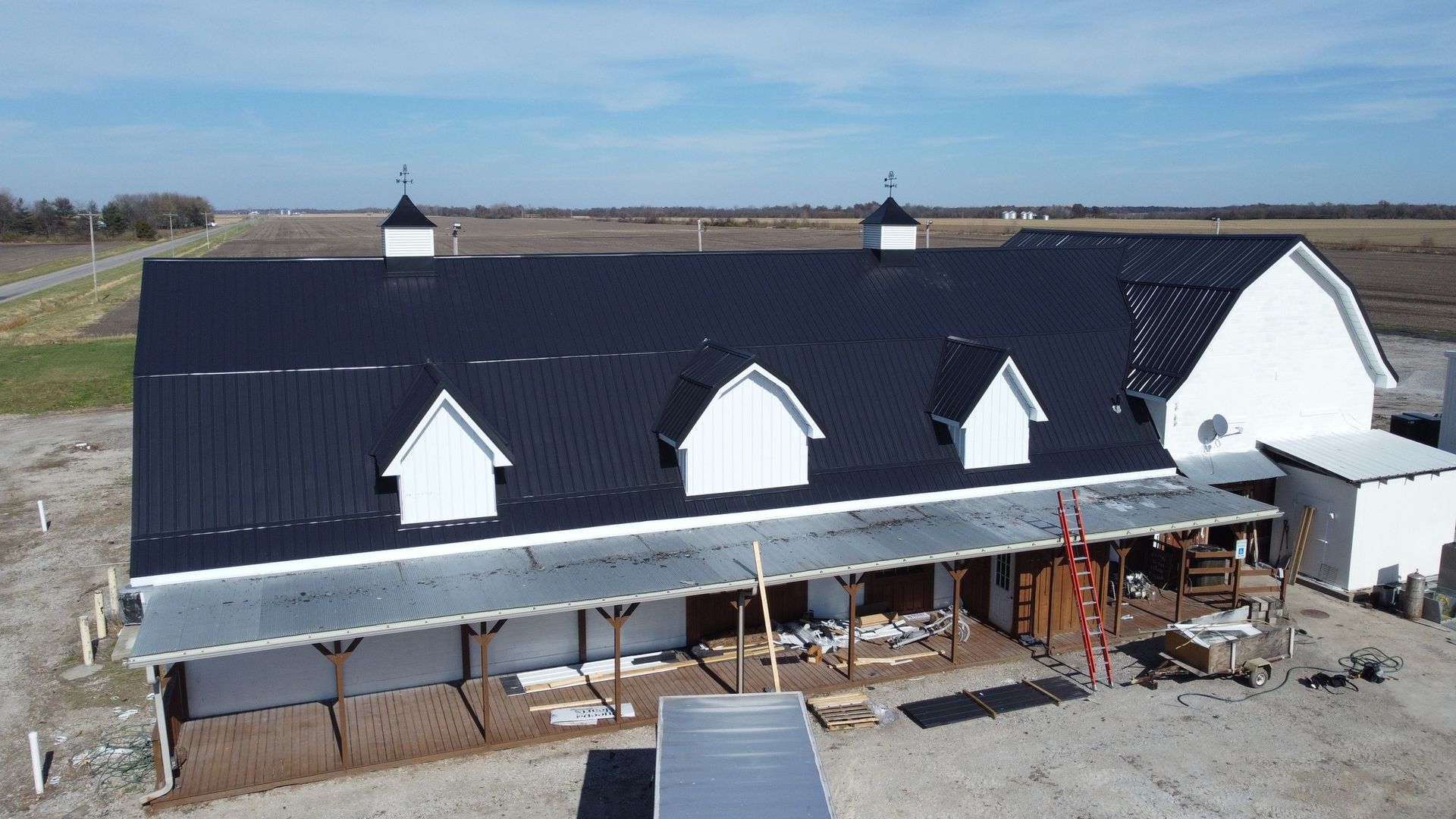 An aerial view of a white, barn-style building with a black metal roof, three dormers, and an unfinished porch area.