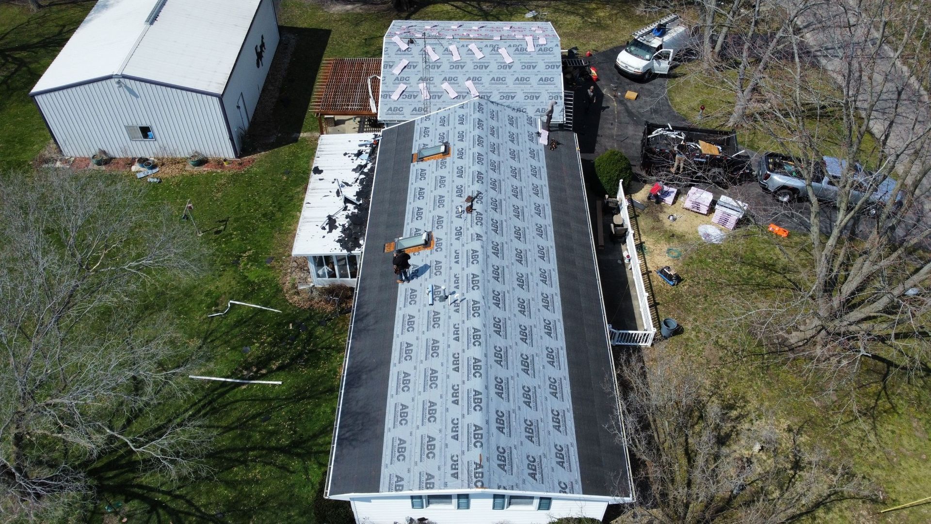 An aerial view shows workers installing new underlayment on a residential roof, with materials and vehicles parked nearby.