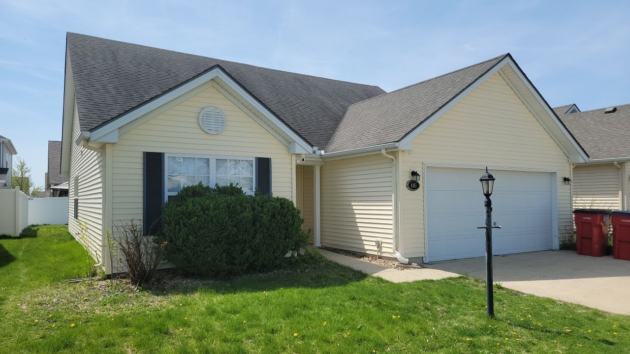 A single-story, light yellow suburban house with a gray shingled roof, a two-car garage, and a manicured front lawn.