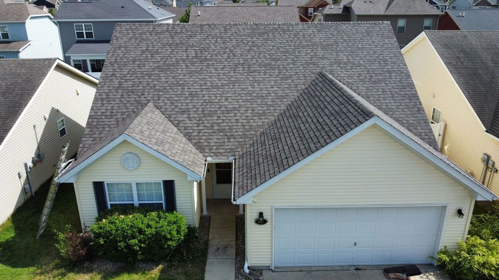 Aerial view of a light yellow suburban house with a gray shingled roof and attached two-car garage