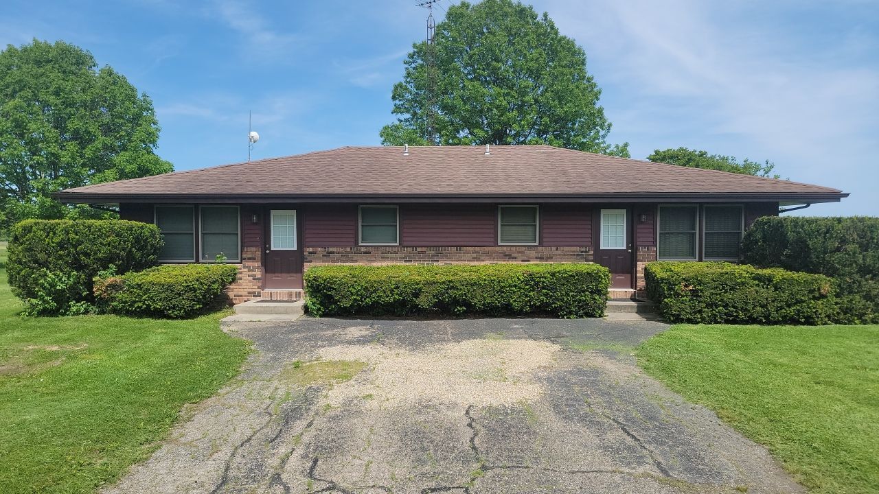 A brown, single-story duplex with a tan brick base, centered front doors, and a gravel driveway, surrounded by greenery.