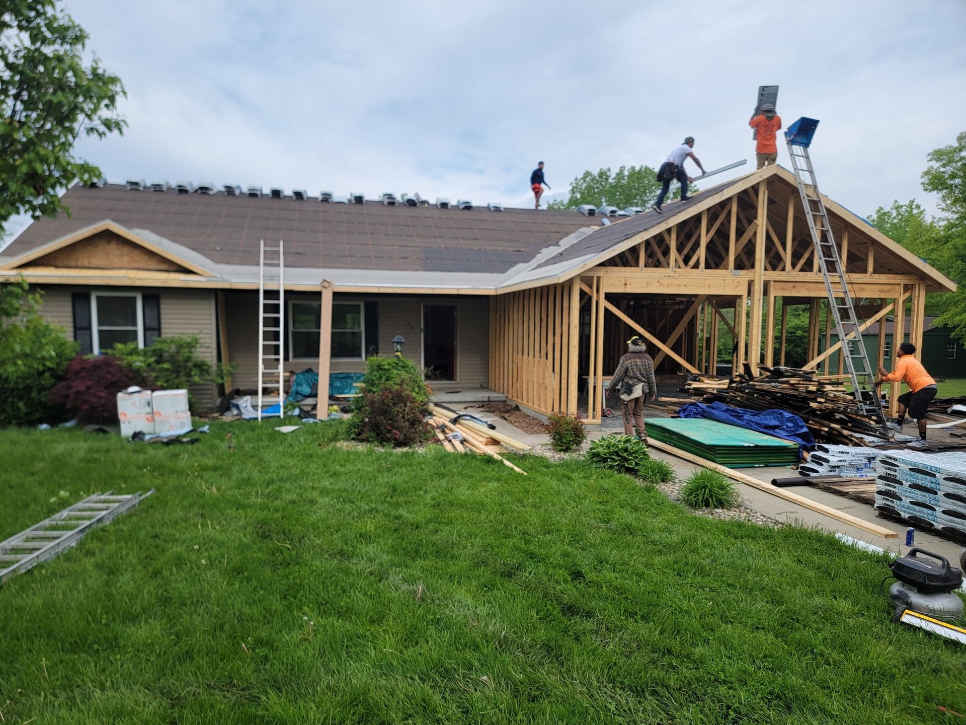 Workers install roofing on a house undergoing construction with an exposed wooden frame extension on a sunny day.