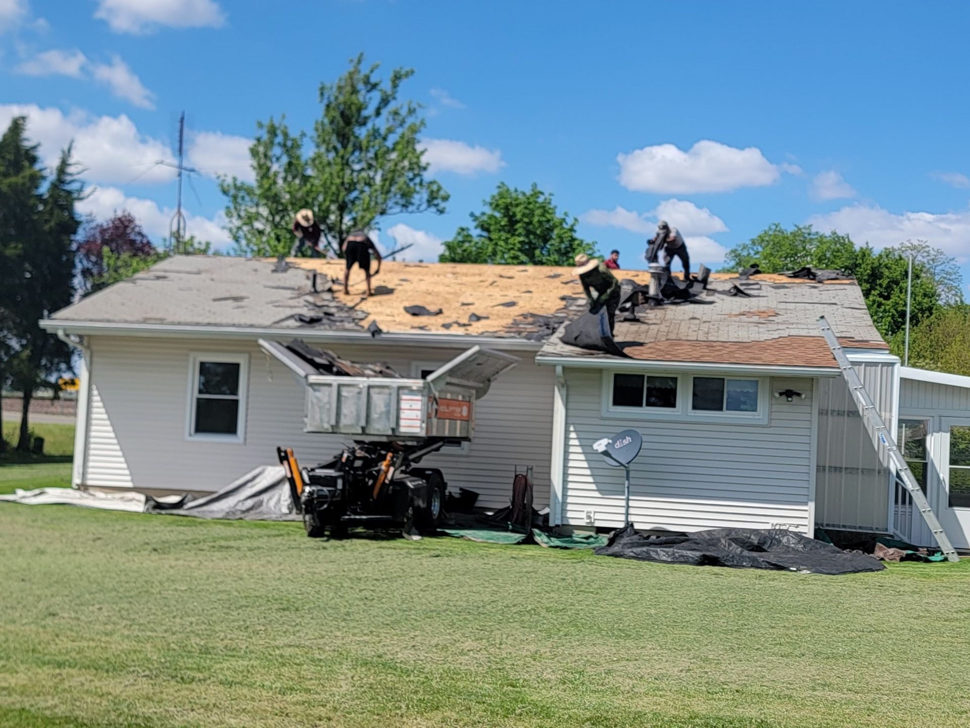 A crew works on a roof replacement on a white house, with a debris lift parked in the yard under a blue sky.