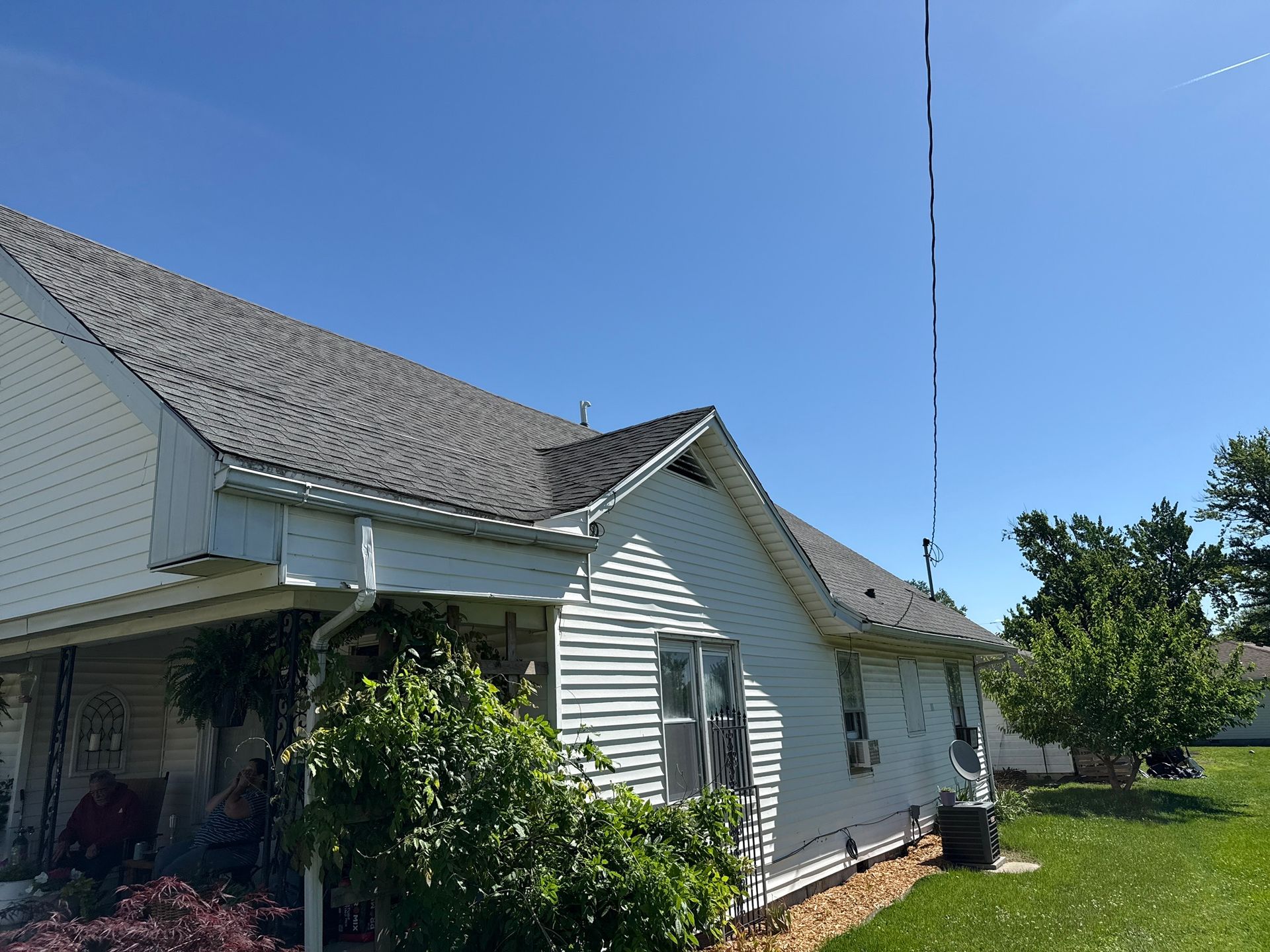 A white, single-story house with a porch and dark shingles under a clear blue sky, featuring a tall, thin antenna mast.