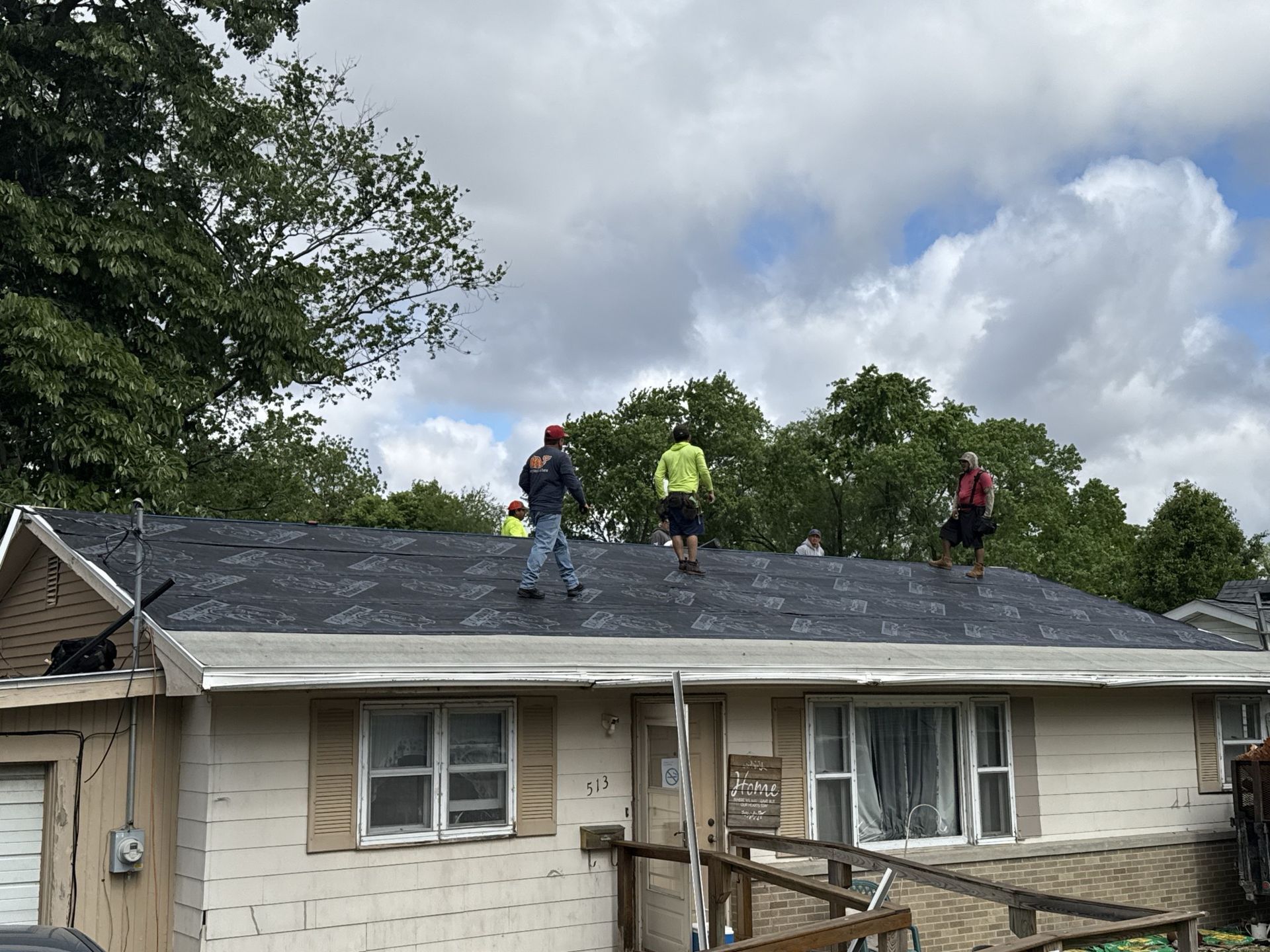 Three workers stand on a residential roof covered with black underlayment, set against a background of trees and sky.