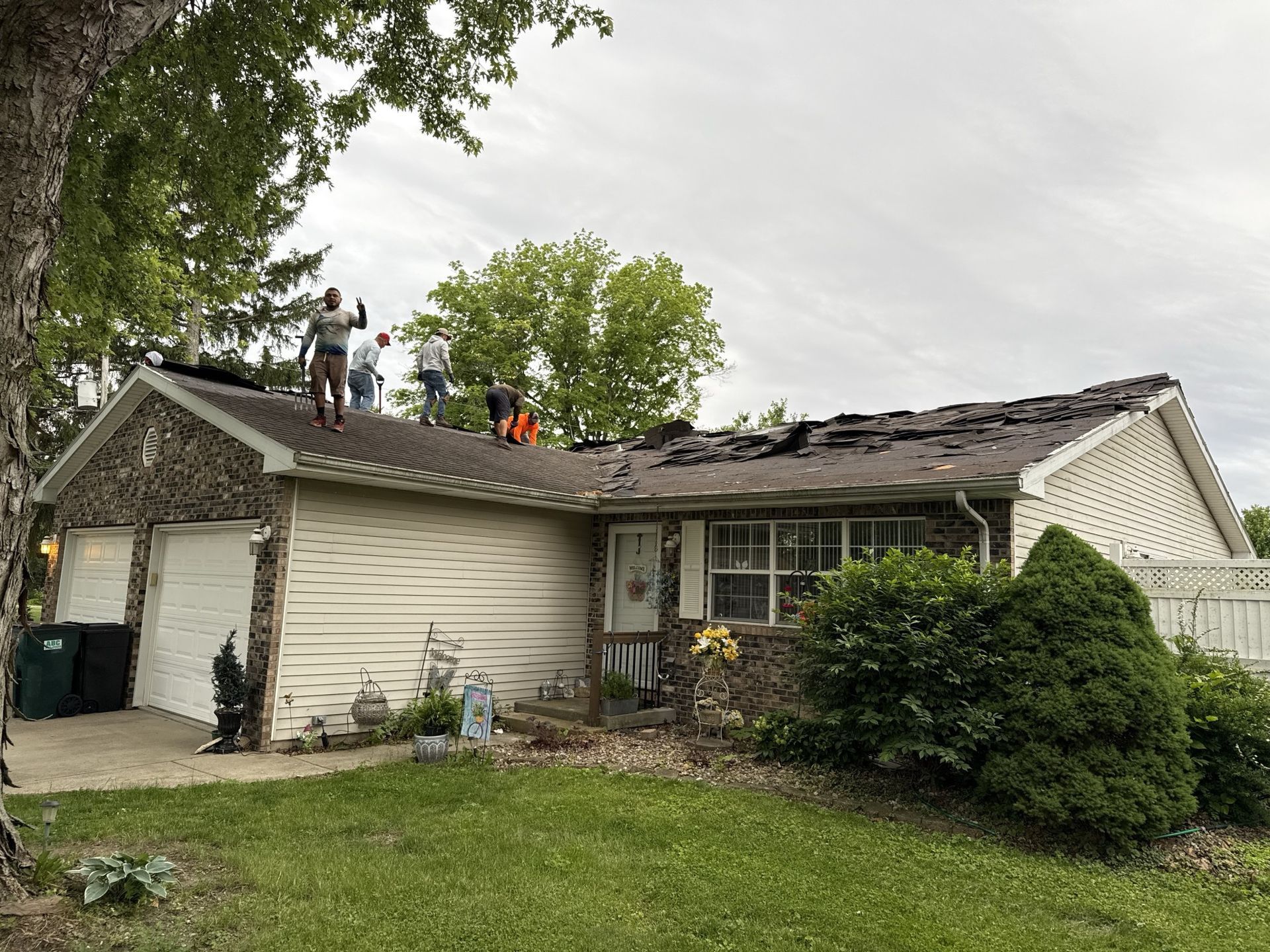 Four workers are removing or replacing shingles on the roof of a beige one-story house with a stone facade.