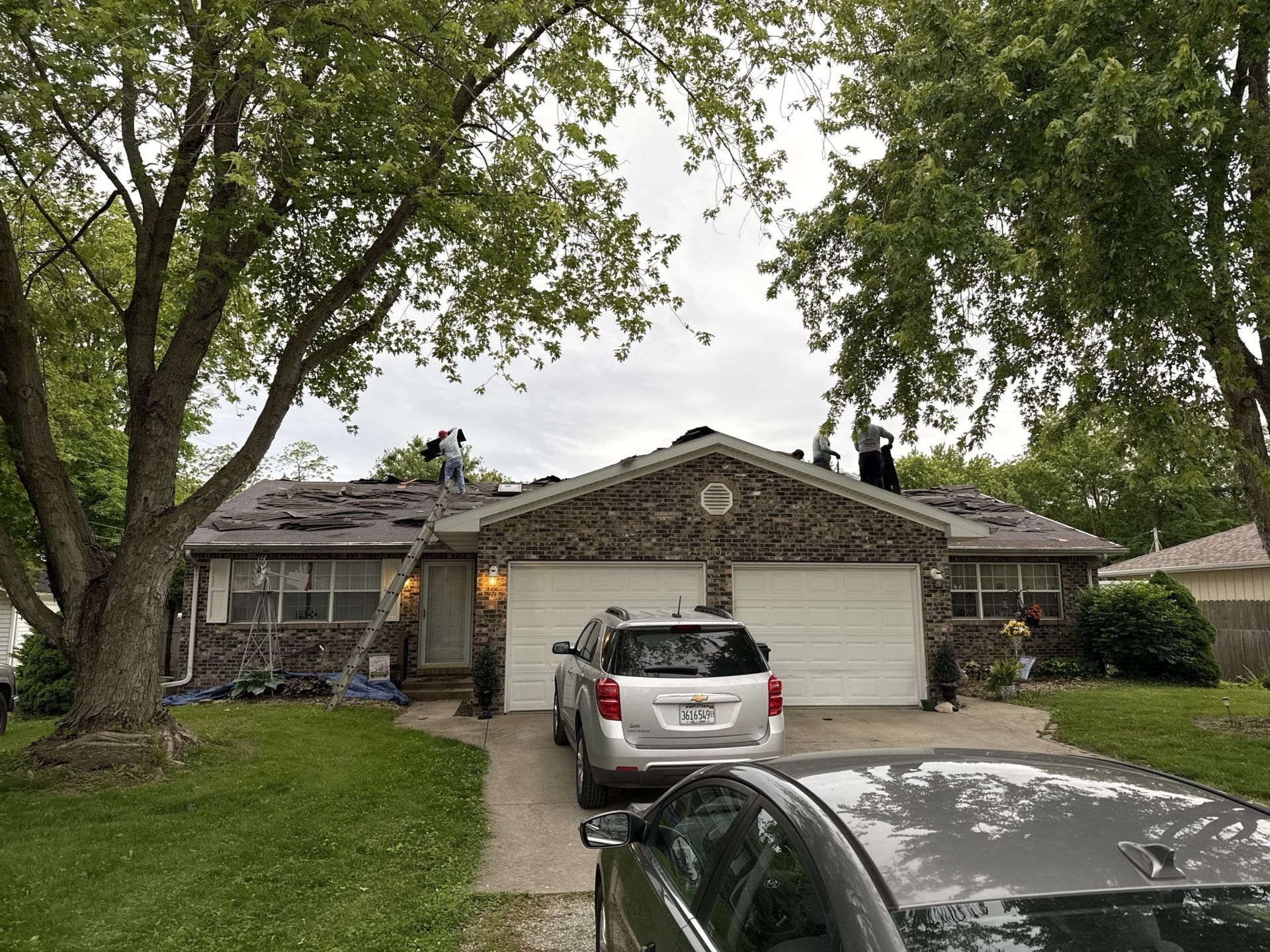 Roofers working on the roof of a suburban stone-faced house with two cars parked in the driveway.