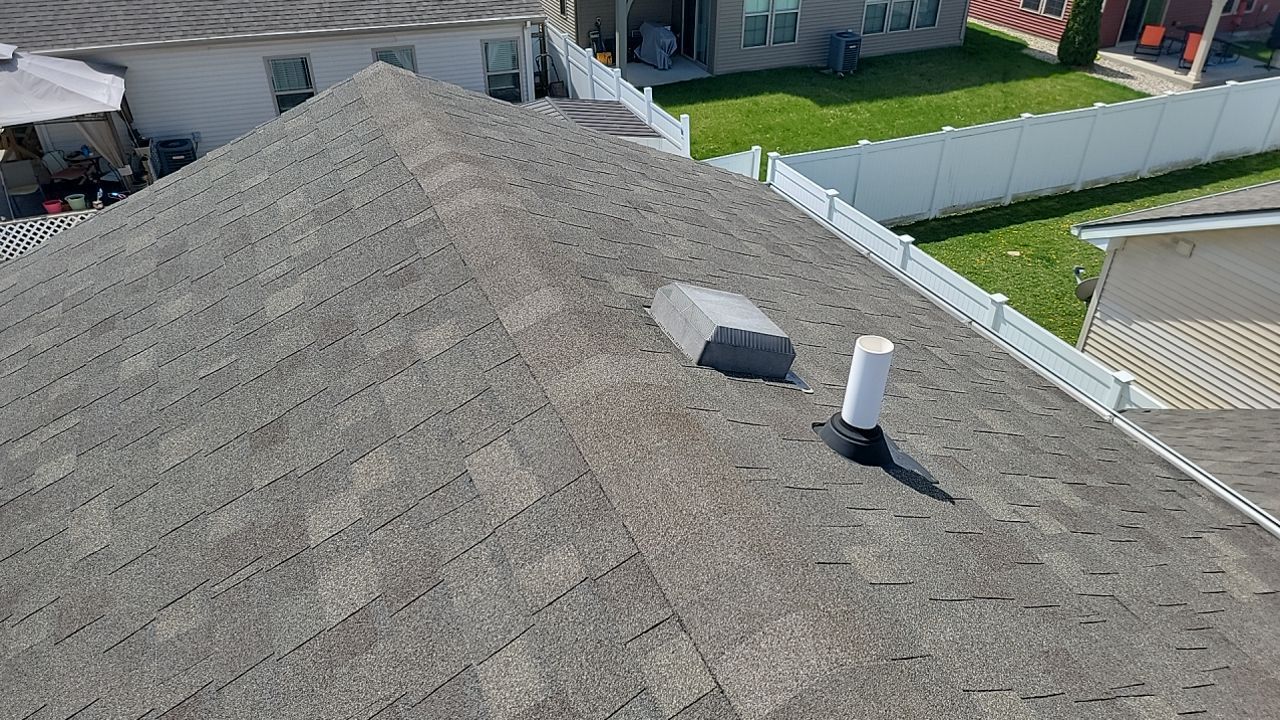 High-angle view of a gray asphalt shingle roof with a vent and a plumbing pipe, viewed from above a white backyard fence.