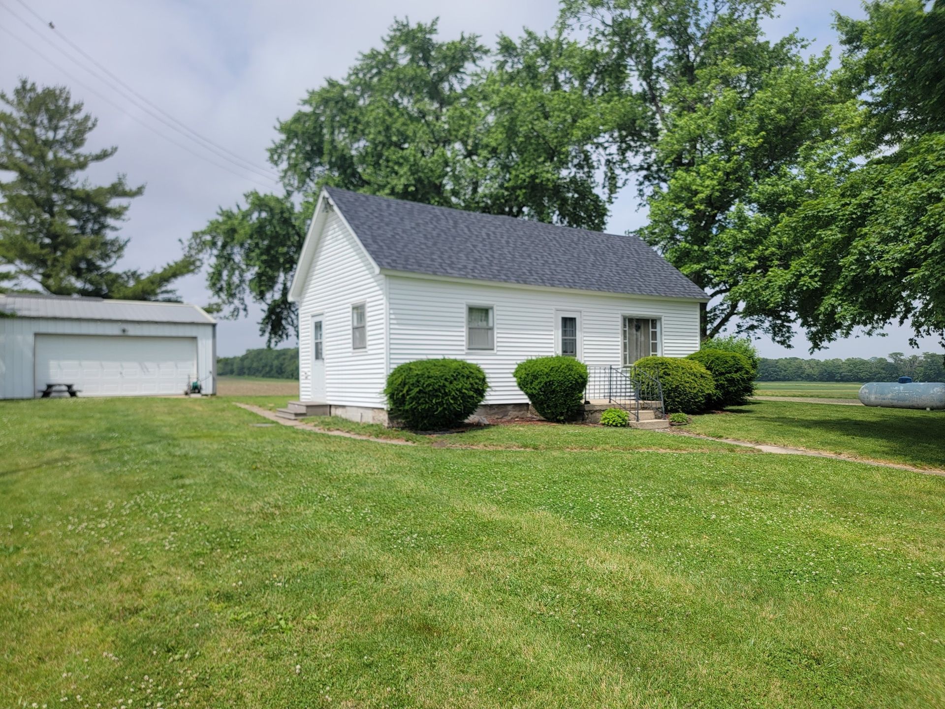 A white, single-story house with a grey roof and a detached garage on a grassy lot surrounded by trees.