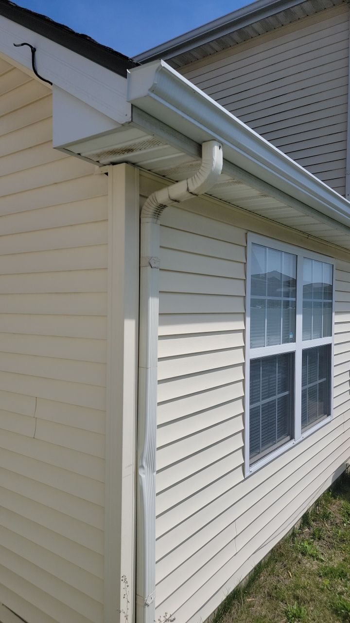 A beige house exterior showing a bent white metal downspout attached to the corner near a window under a roof gutter.