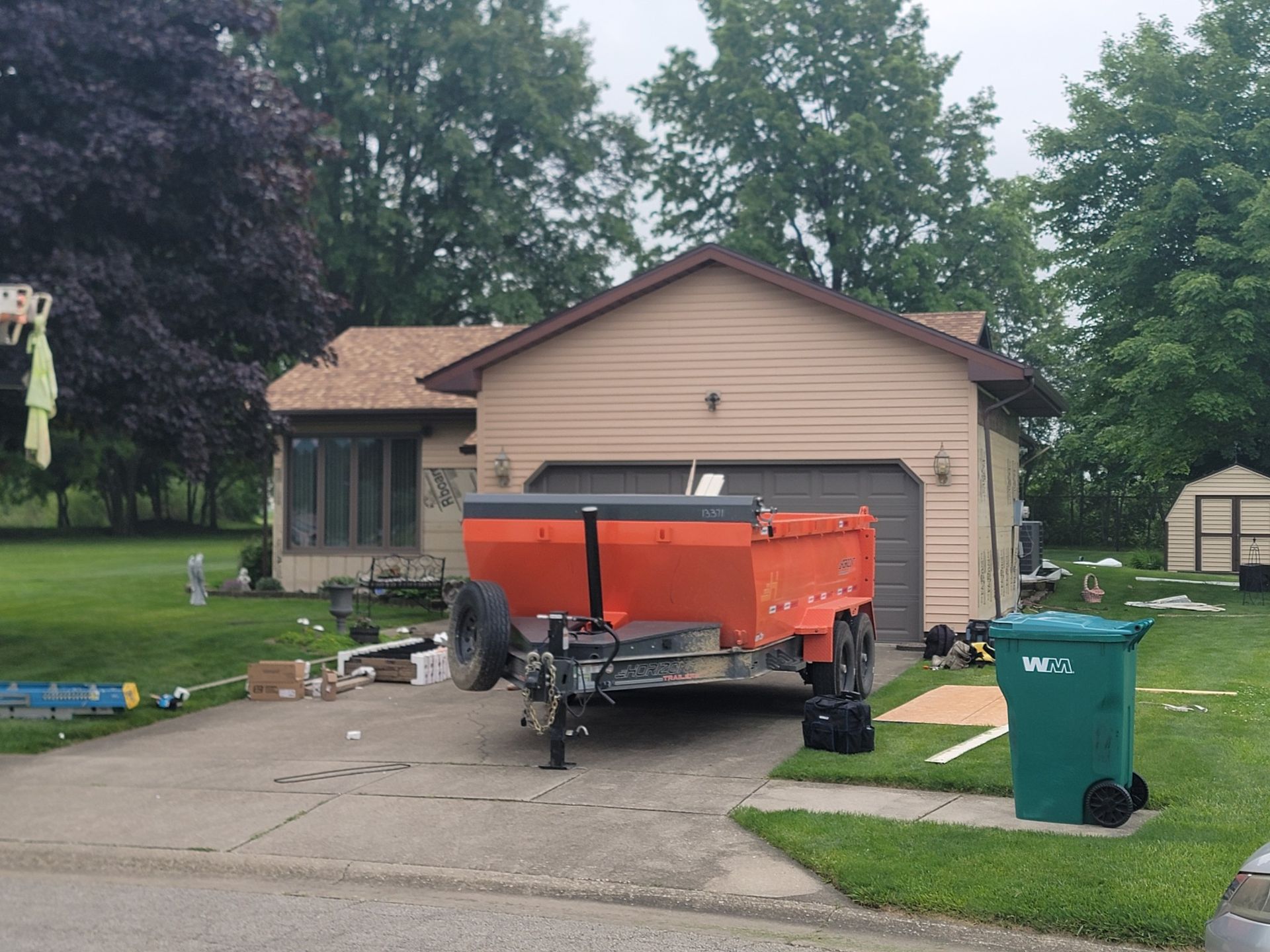 An orange dump trailer sits on a concrete driveway in front of a house with a tan garage, next to a green trash bin.