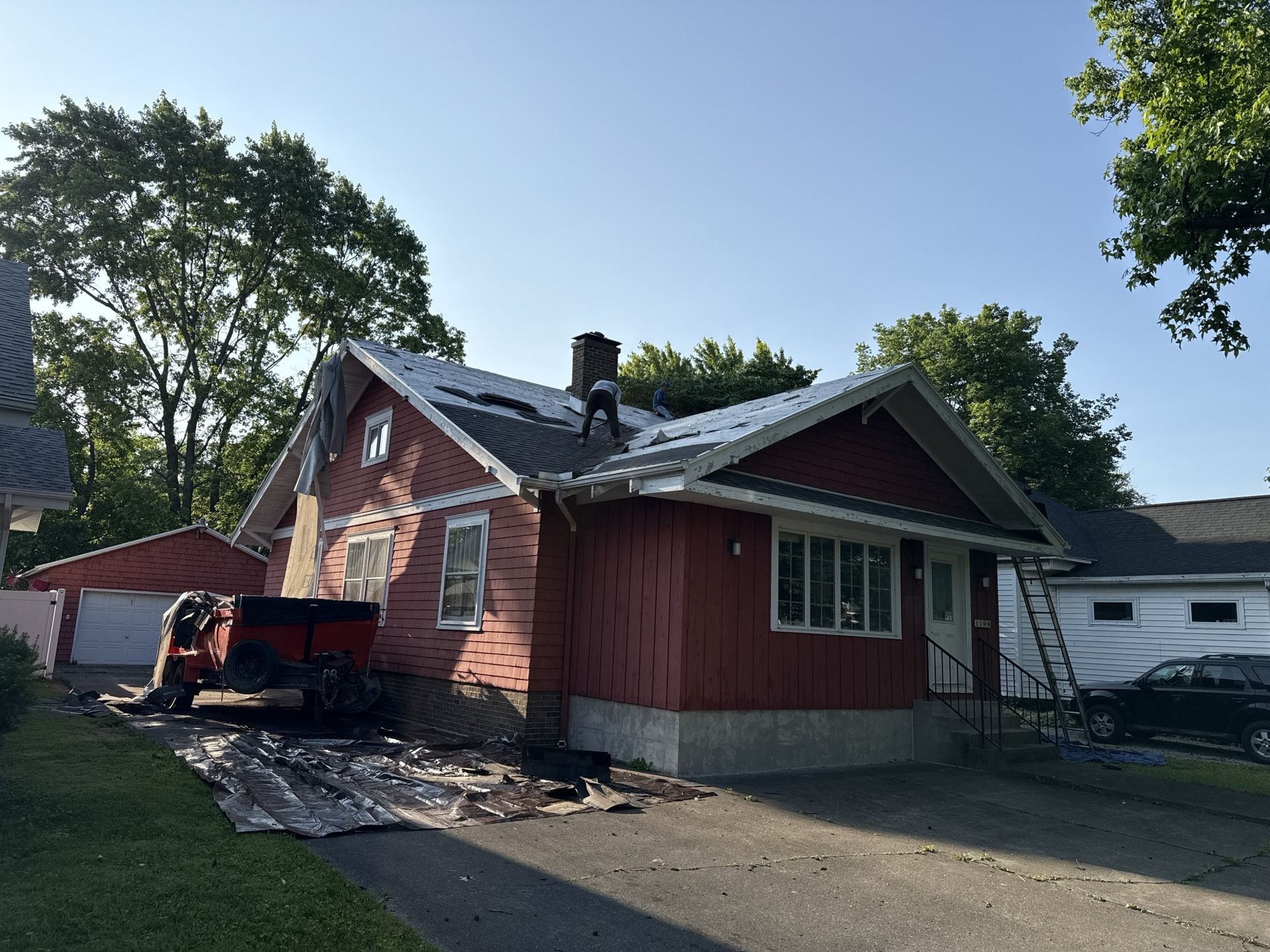 Workers replace the roof of a red single-story house with a chimney, featuring a dumpster and ladder on site.