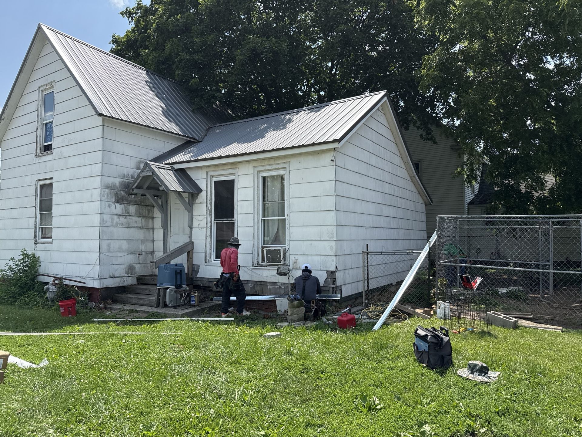 Two people work on the exterior of a white, two-story house with a metal roof on a sunny day.