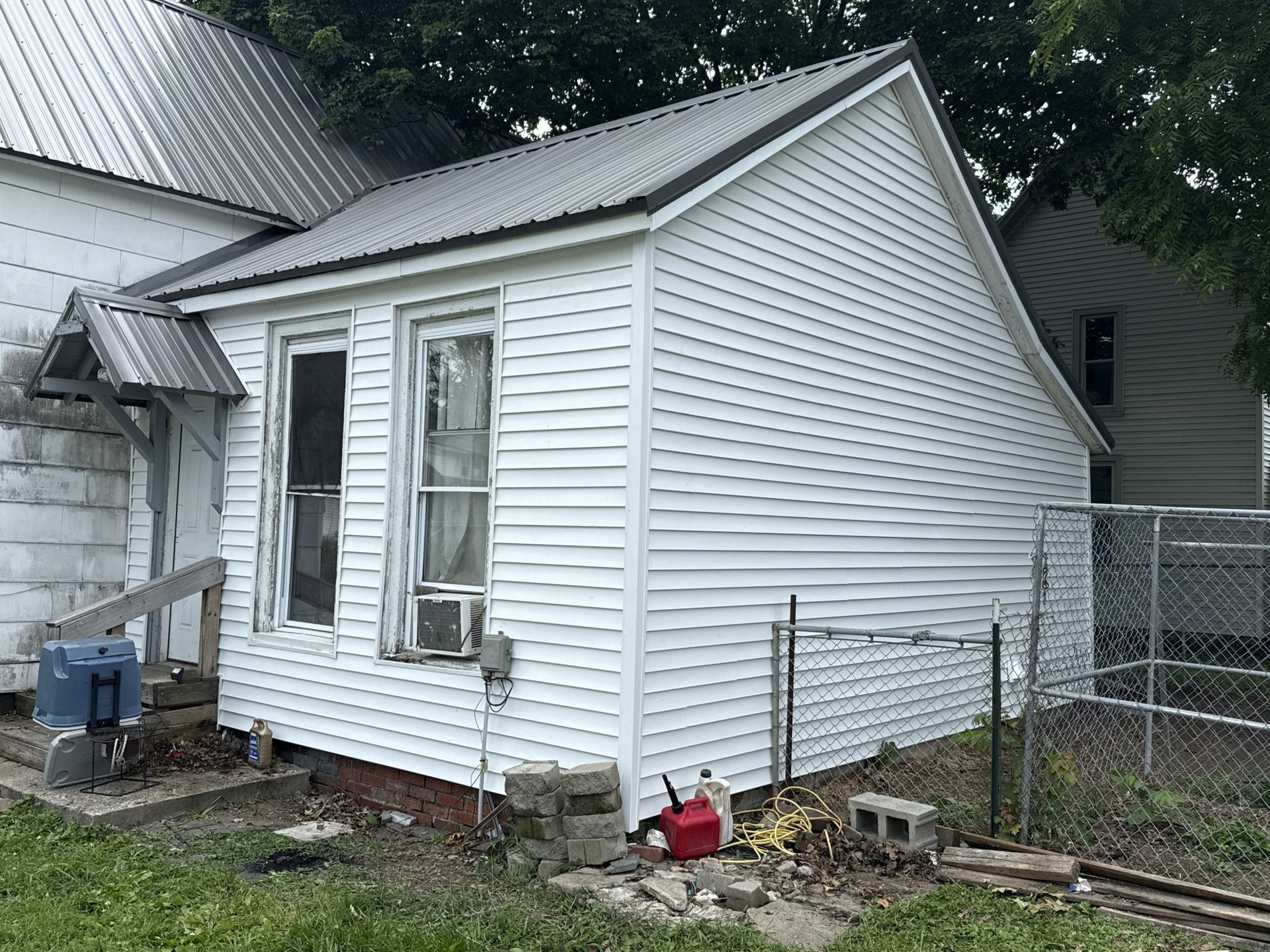 A small, white, single-story house with a metal roof, located in a yard with a chain-link fence.