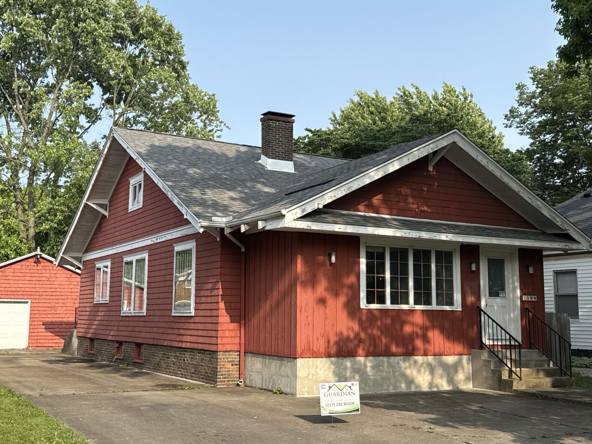 A single-story red house with a dark roof and chimney, situated on a paved lot with a detached red garage.