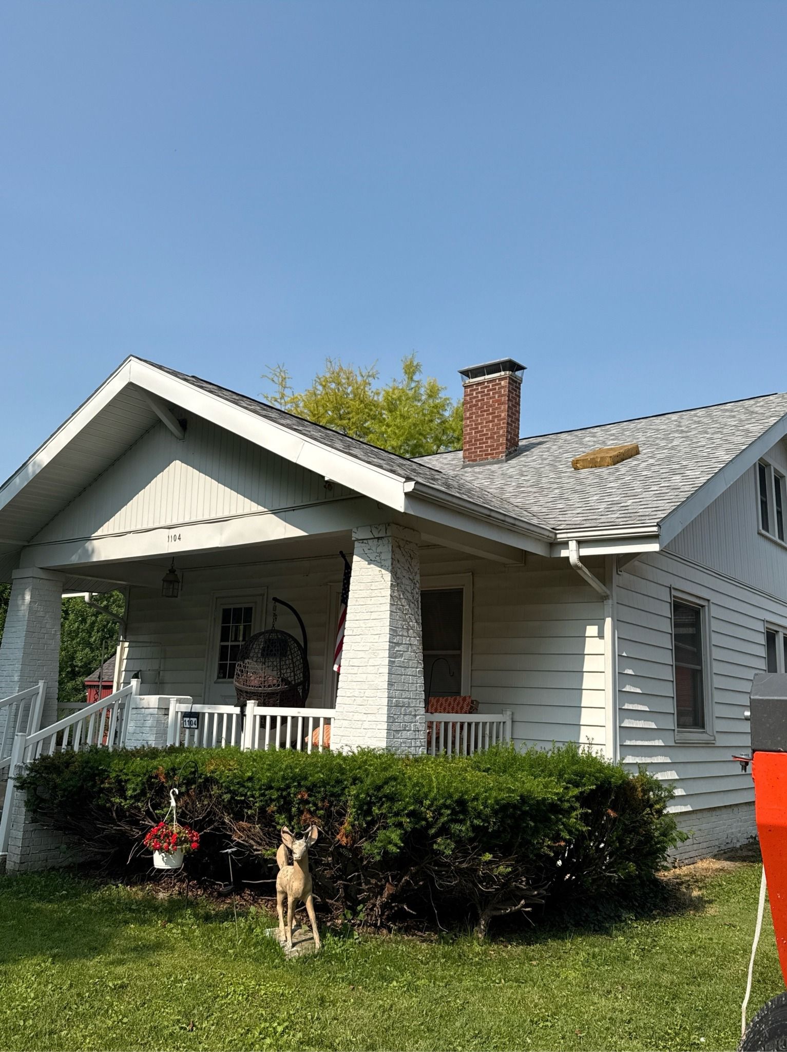 A small, white, single-story house with a brick chimney and a front porch, viewed from a grassy yard under a blue sky.