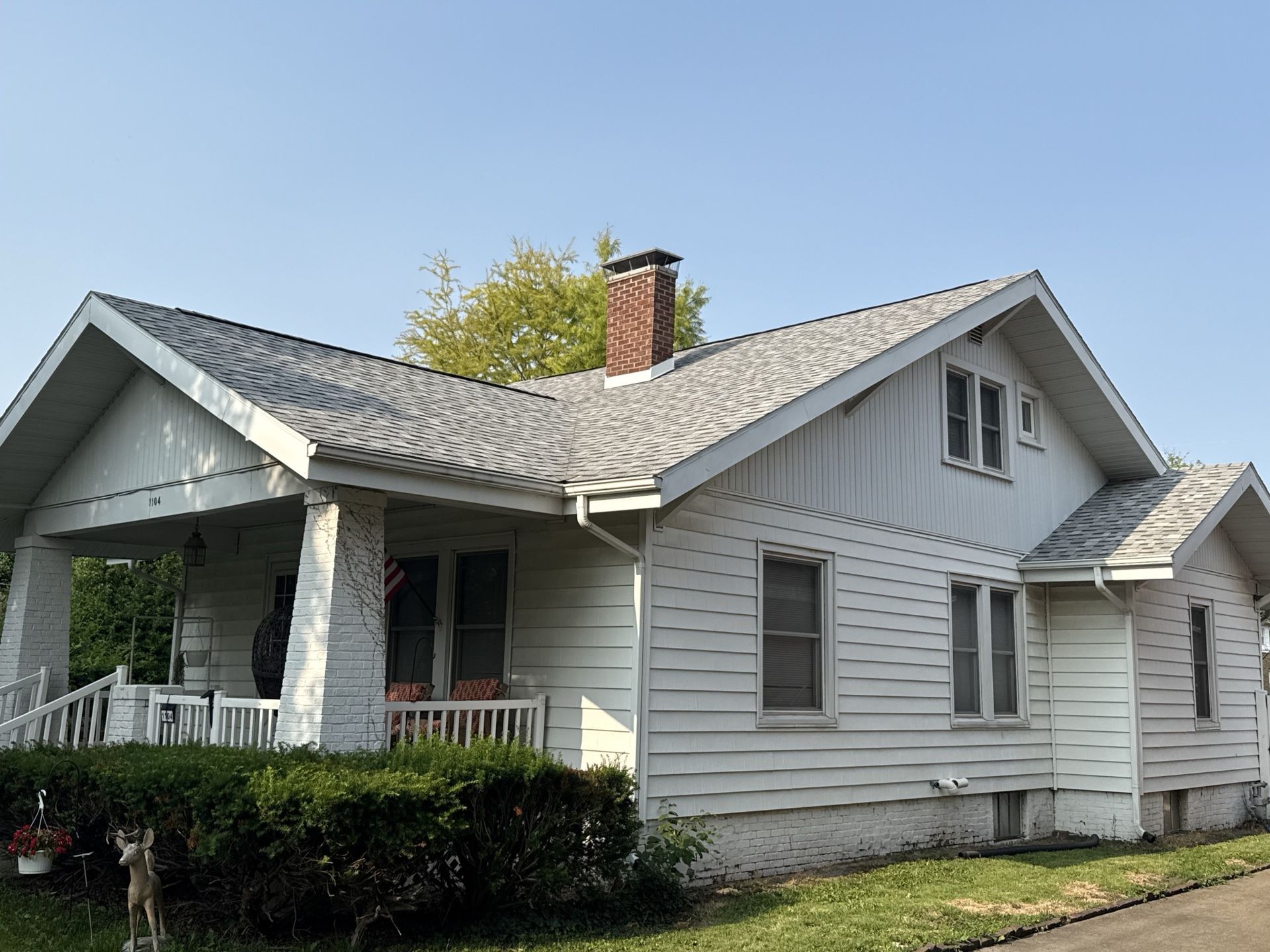 A single-story white house with a brick chimney, front porch, and shingled roof under a clear blue sky.