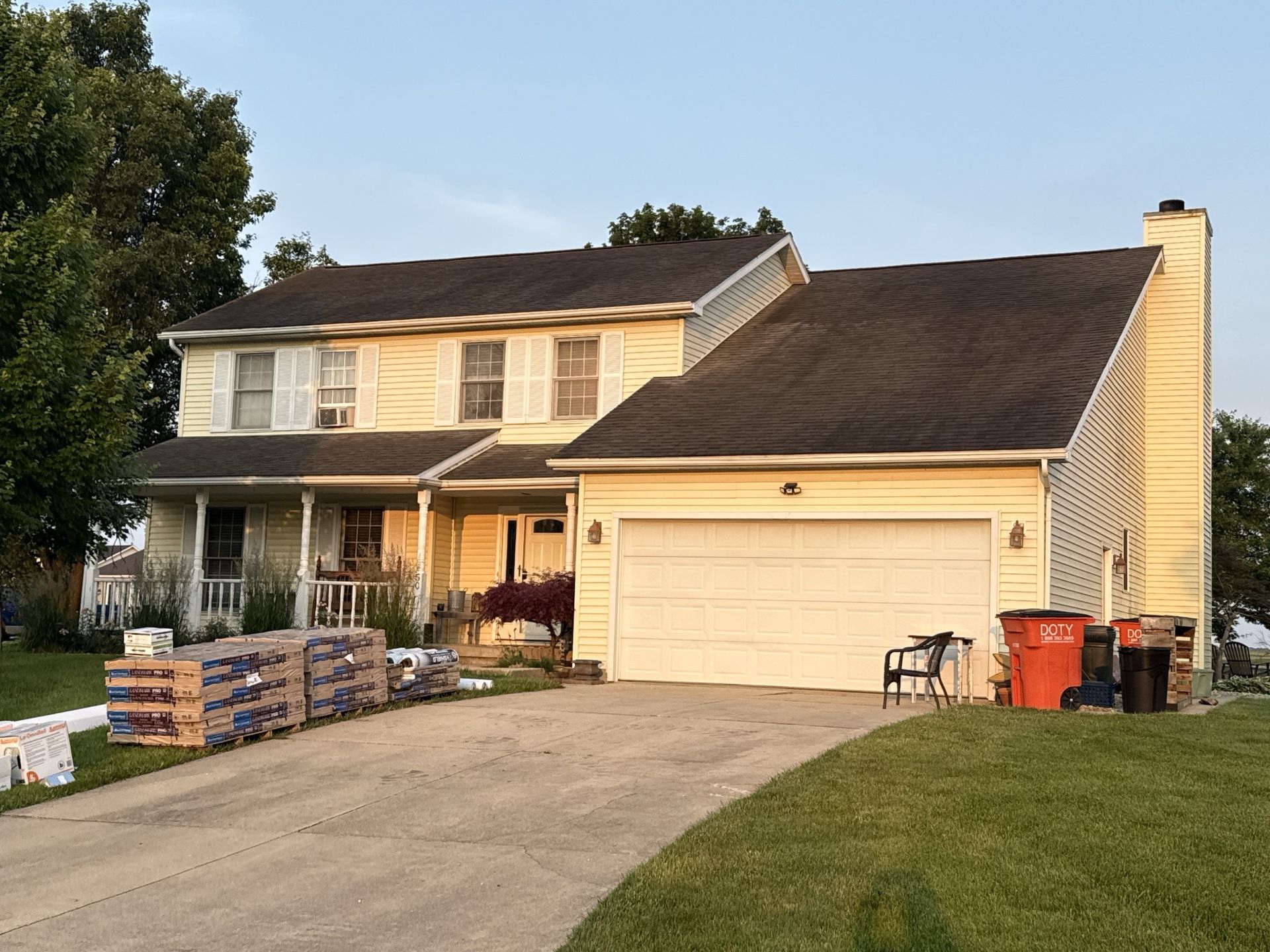 A yellow two-story house with a white garage, a front porch, and stacks of construction materials on the driveway.