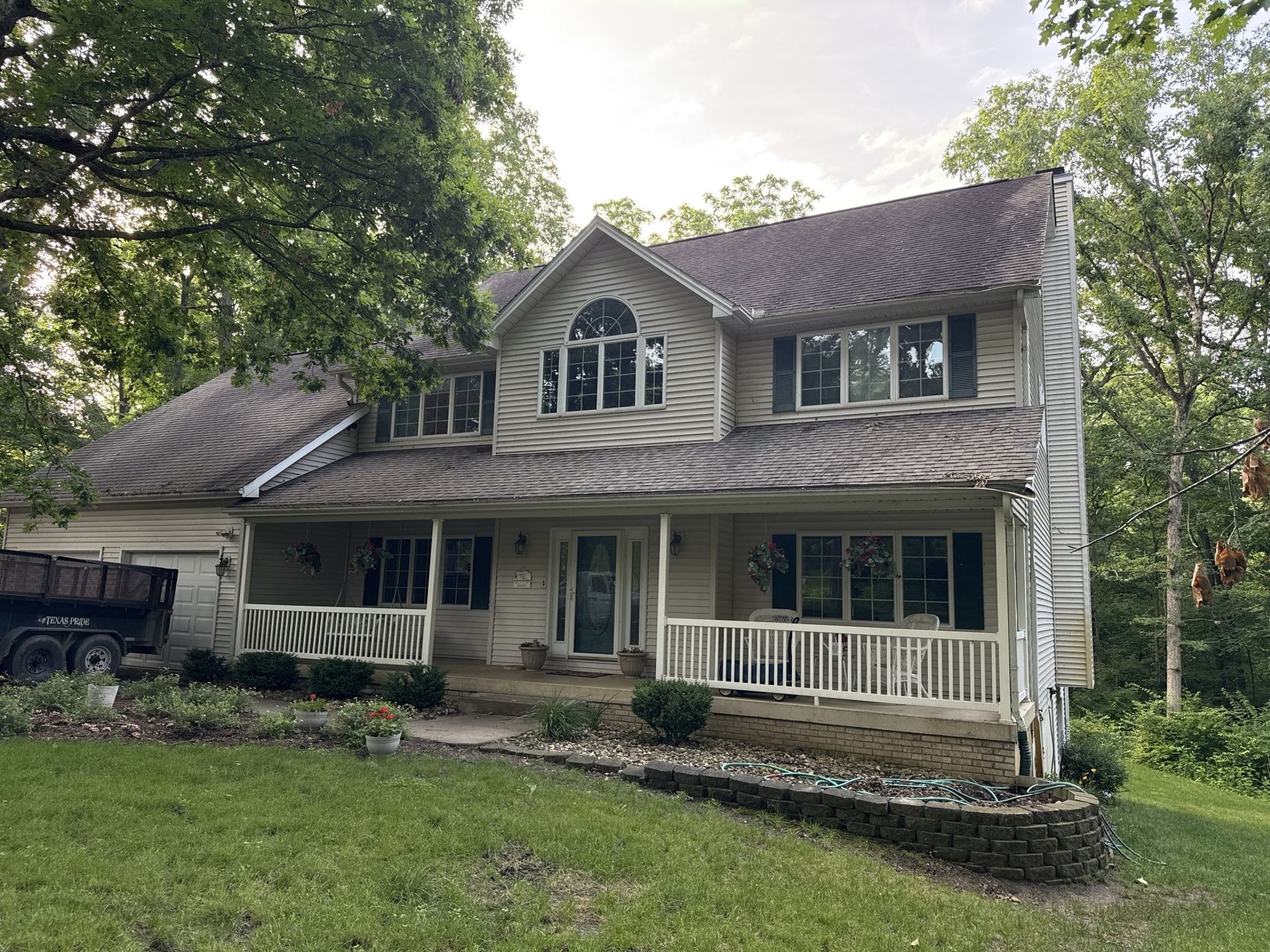 A two-story, light-colored suburban house with a wraparound porch, front gable, and dark roof, surrounded by trees.