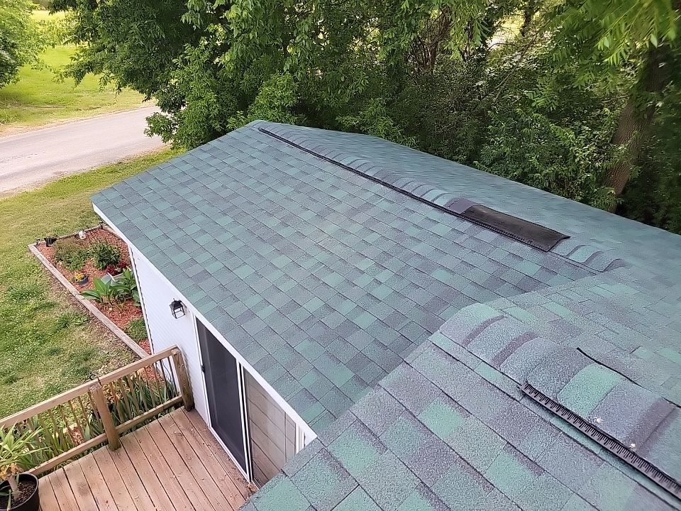 High-angle view of a green asphalt shingle roof on a small building, with a deck and trees in the background.