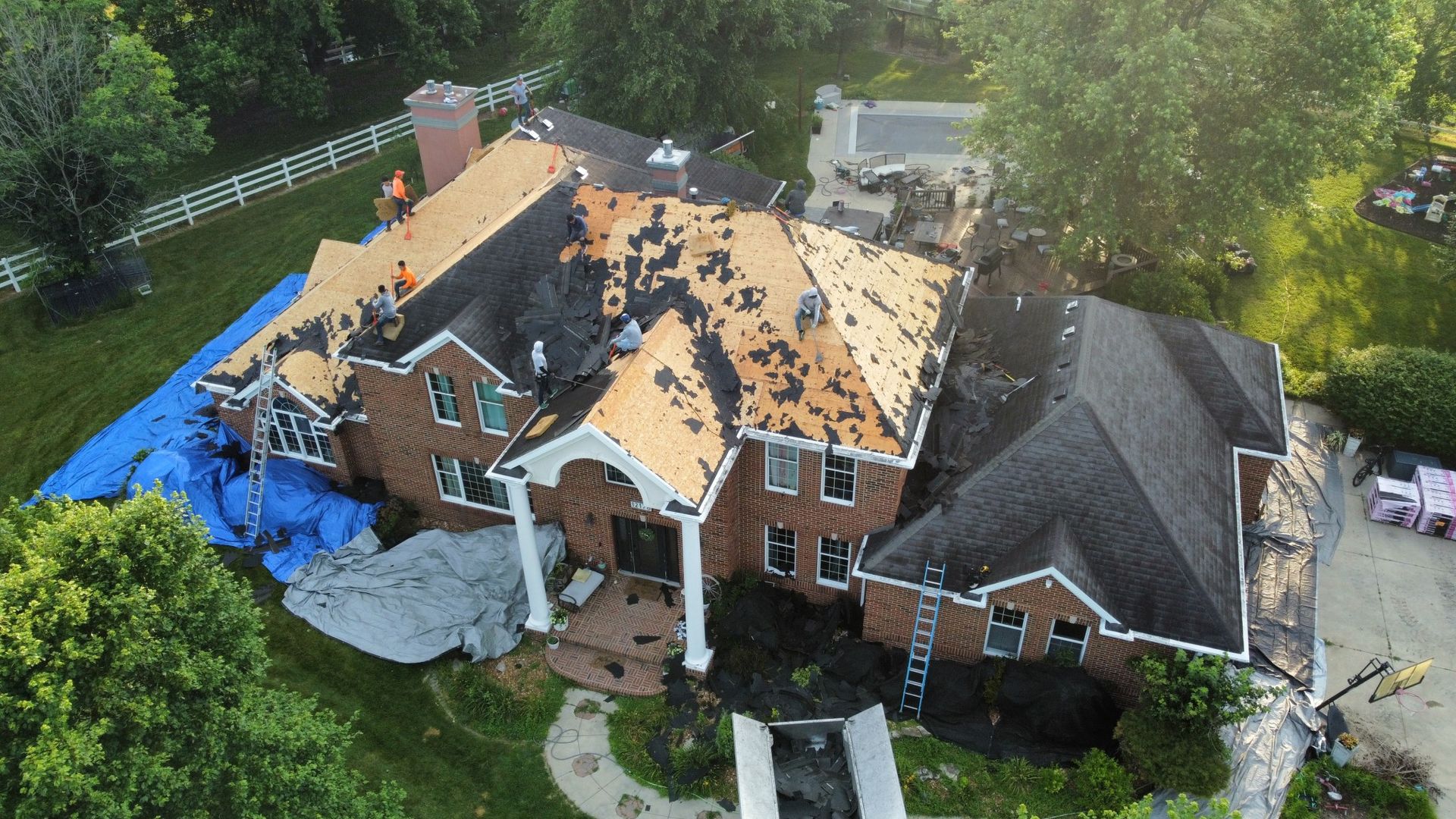 Aerial view of a large brick house undergoing roof repairs, with workers visible on sections of exposed roof decking.