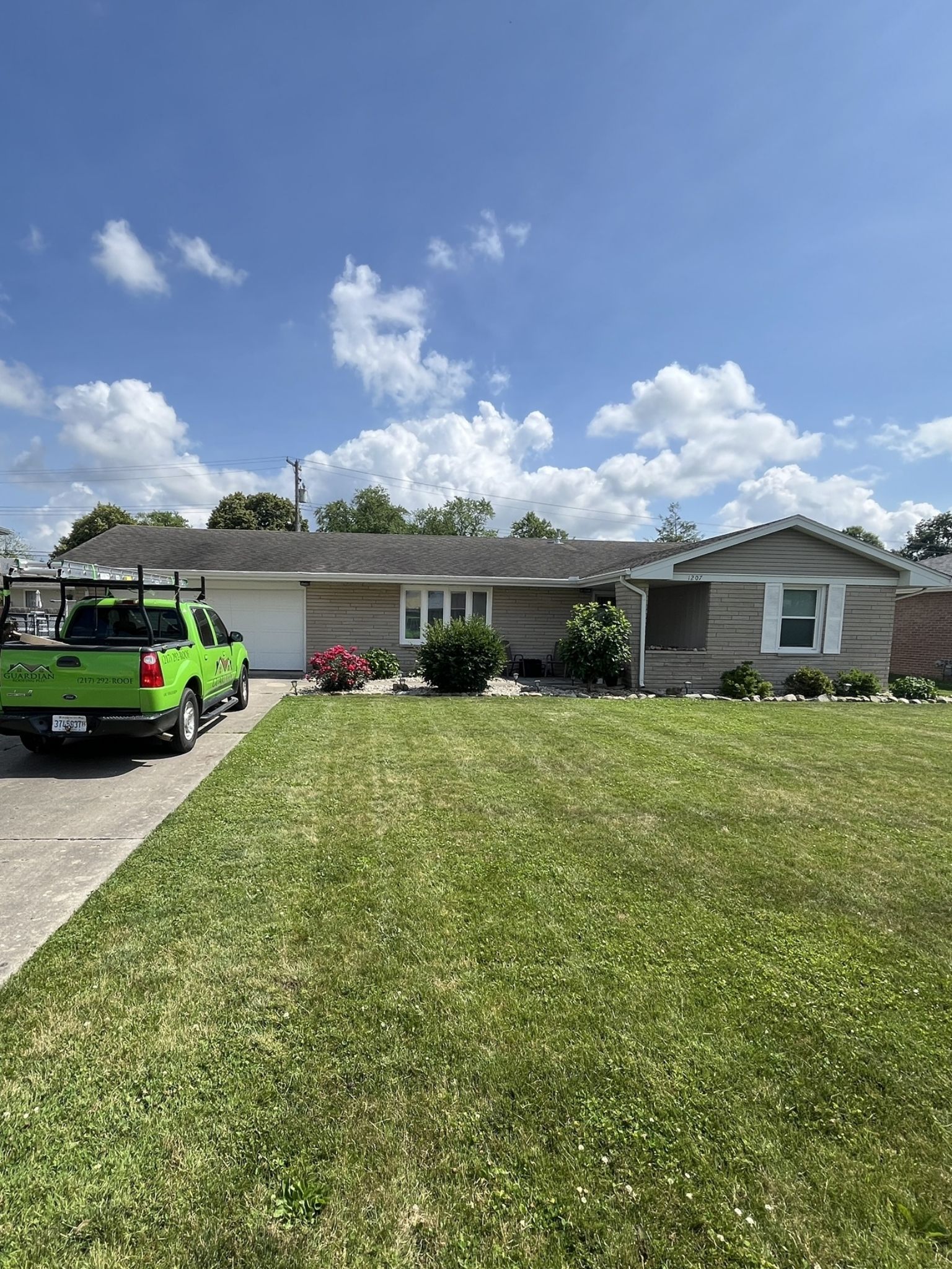 A light gray, single-story house with a green pickup truck in the driveway under a bright blue sky with white clouds.