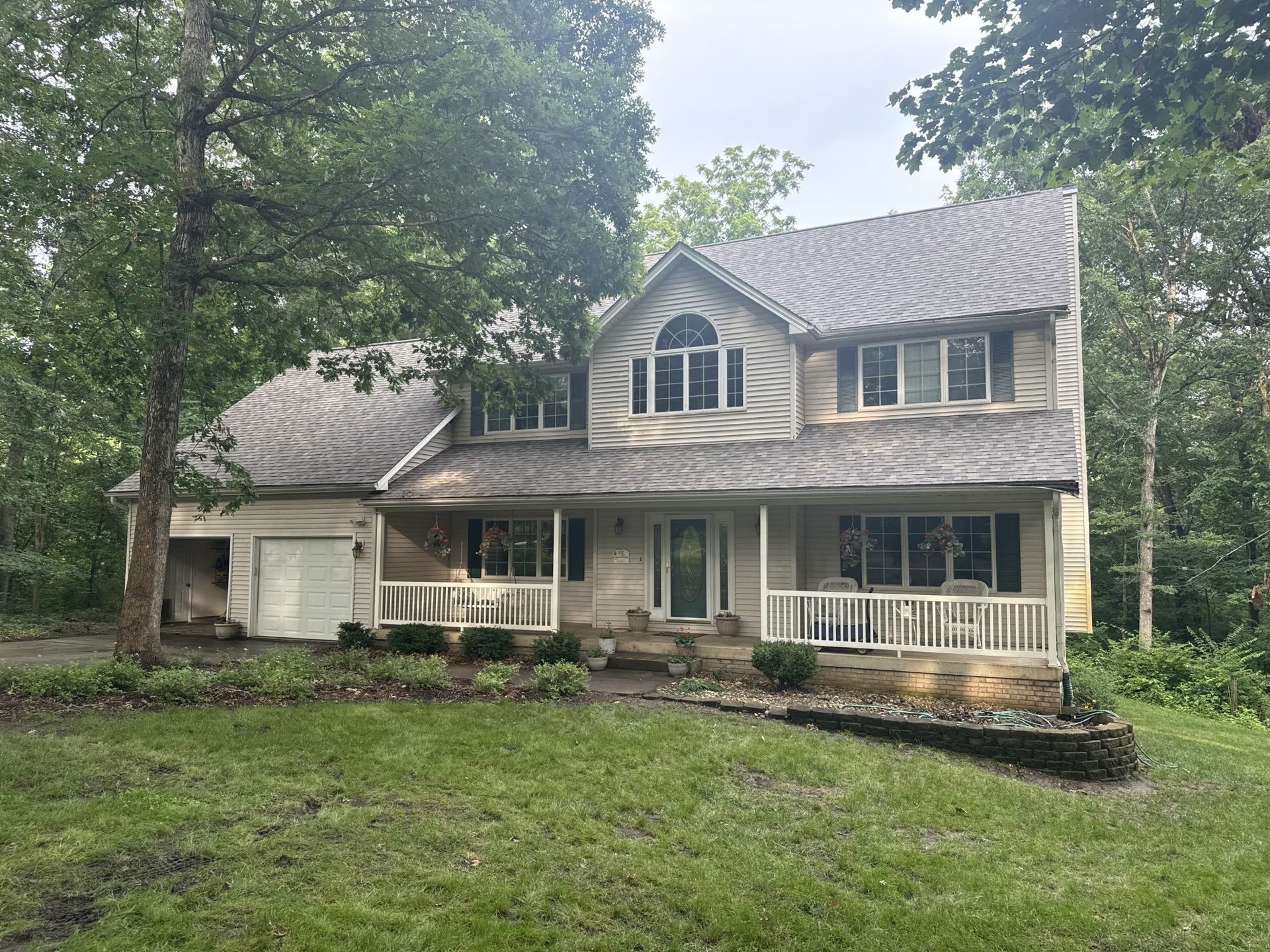 A two-story, light-colored suburban house with a front porch, garage, and arched window, surrounded by lush green trees.