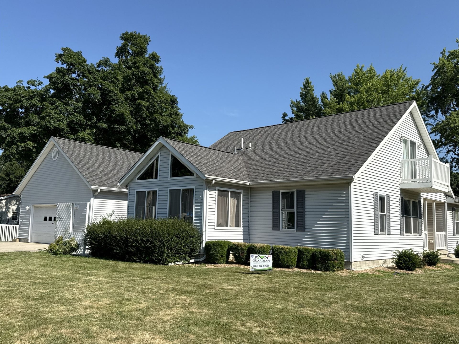 A white, one-story house with a detached garage, a shingled roof, and a small balcony, set against a clear blue sky.