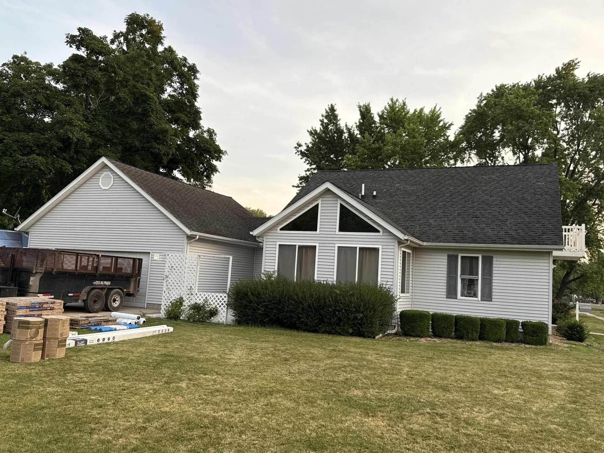 A grey house with a dark roof and large windows stands next to a detached garage with construction supplies nearby.